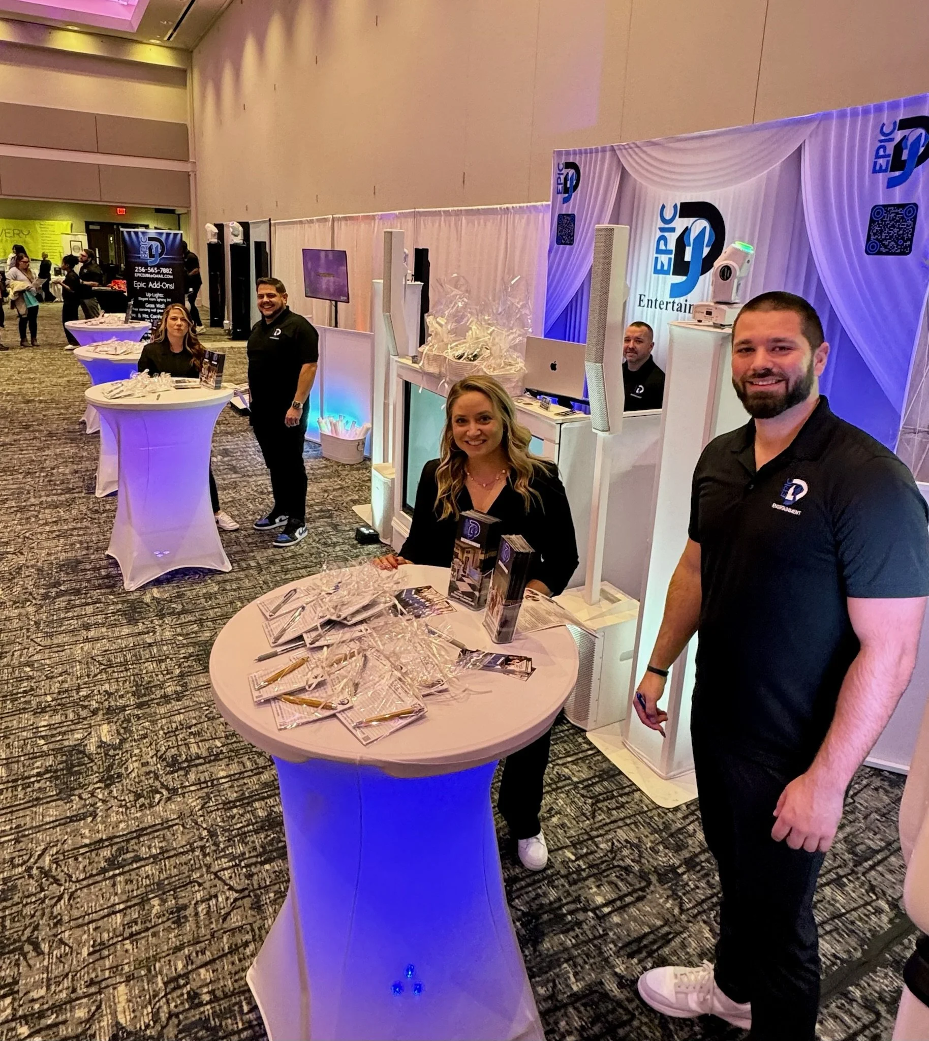 People at a trade show booth with display screens, promotional materials, and branded signage, smiling and greeting attendees.