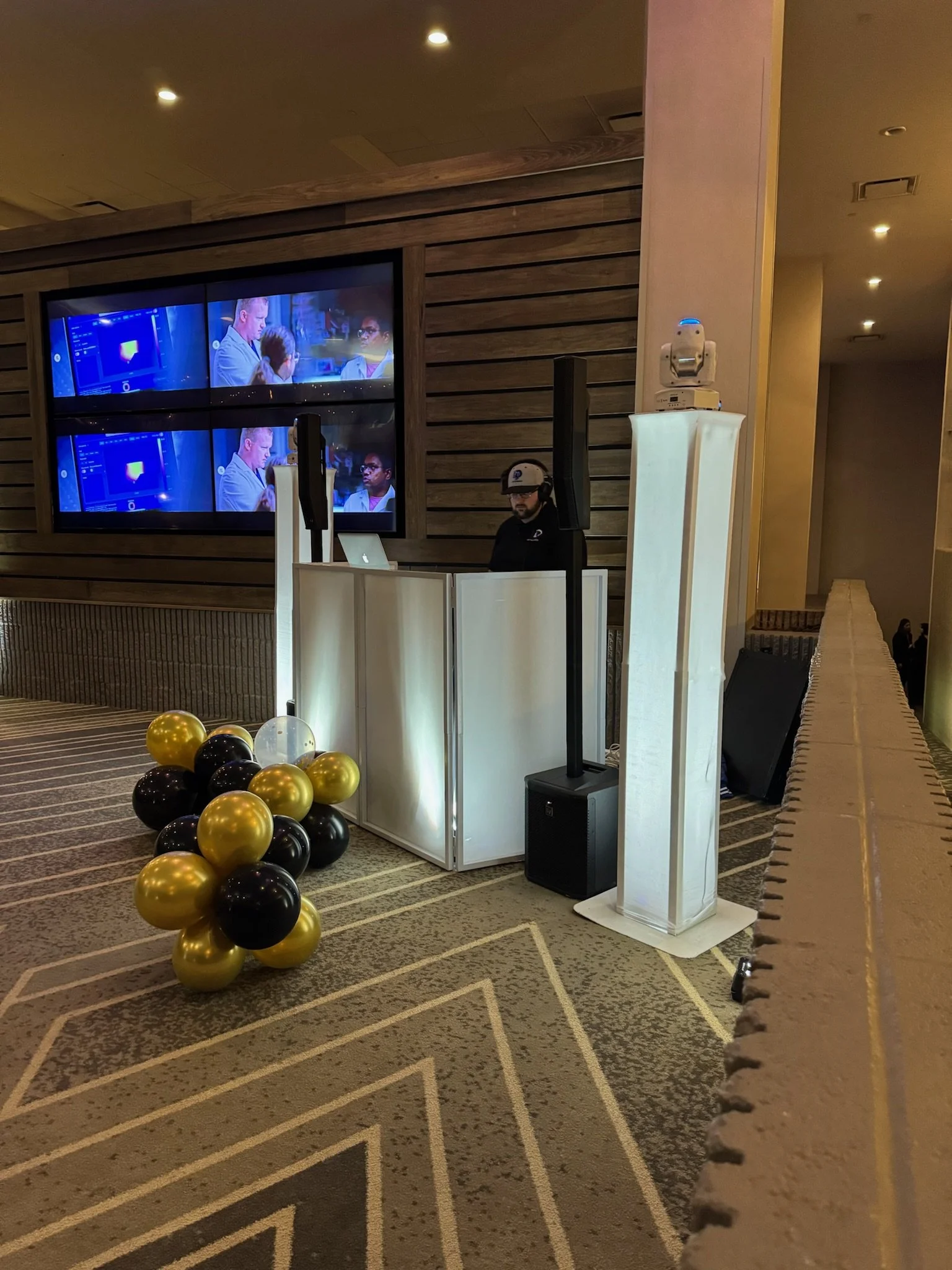 A DJ wearing a hat and headphones behind a white DJ booth with a MacBook, set up near a wall with four screens displaying a TV show with a man and young girl, in a hotel or event space decorated with black and gold balloons.