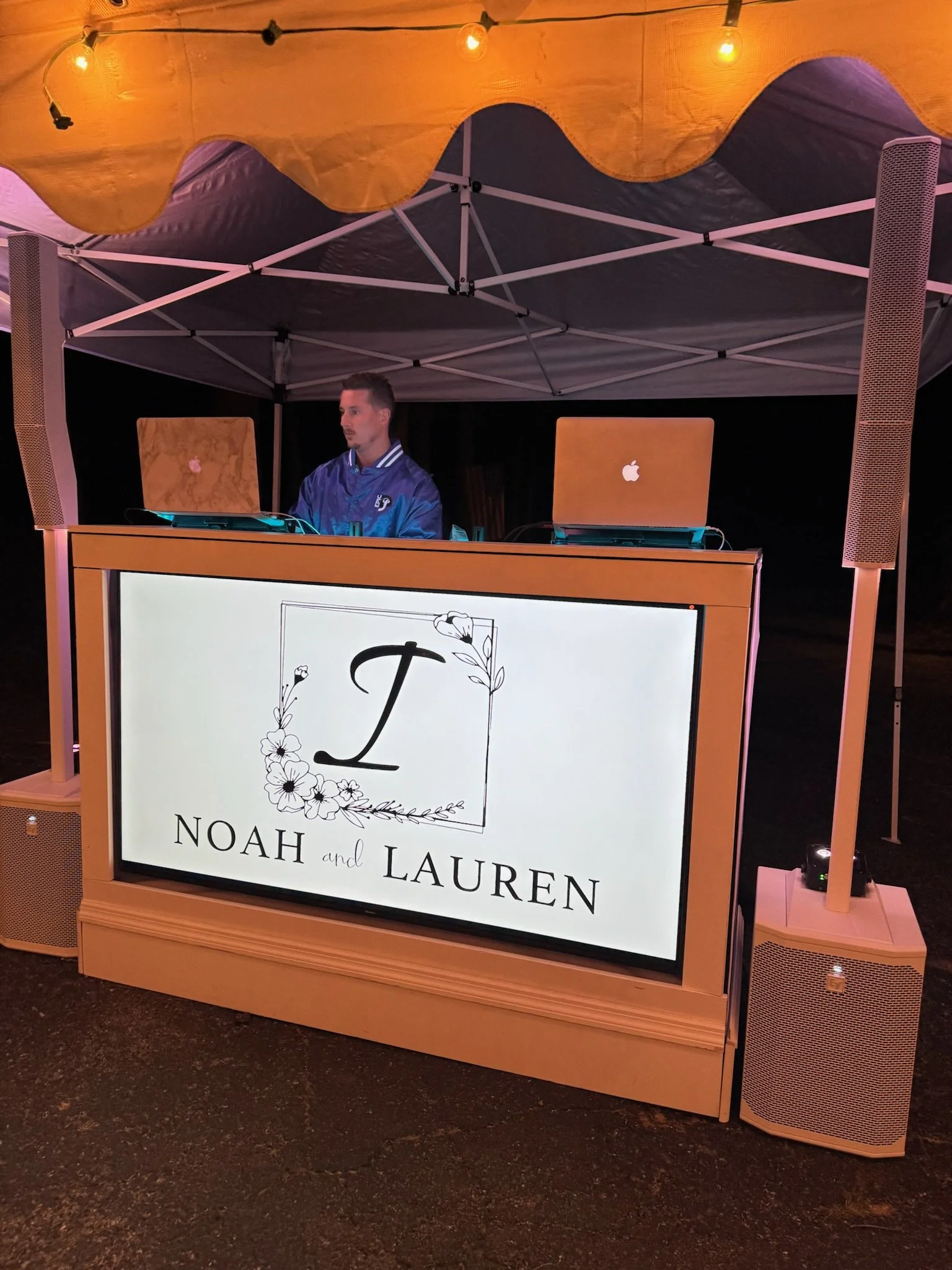 A DJ booth with two laptops, a man in a blue satin jacket, under a canopy with string lights. The booth has a sign that reads 'Noah and Lauren' with floral decorations.