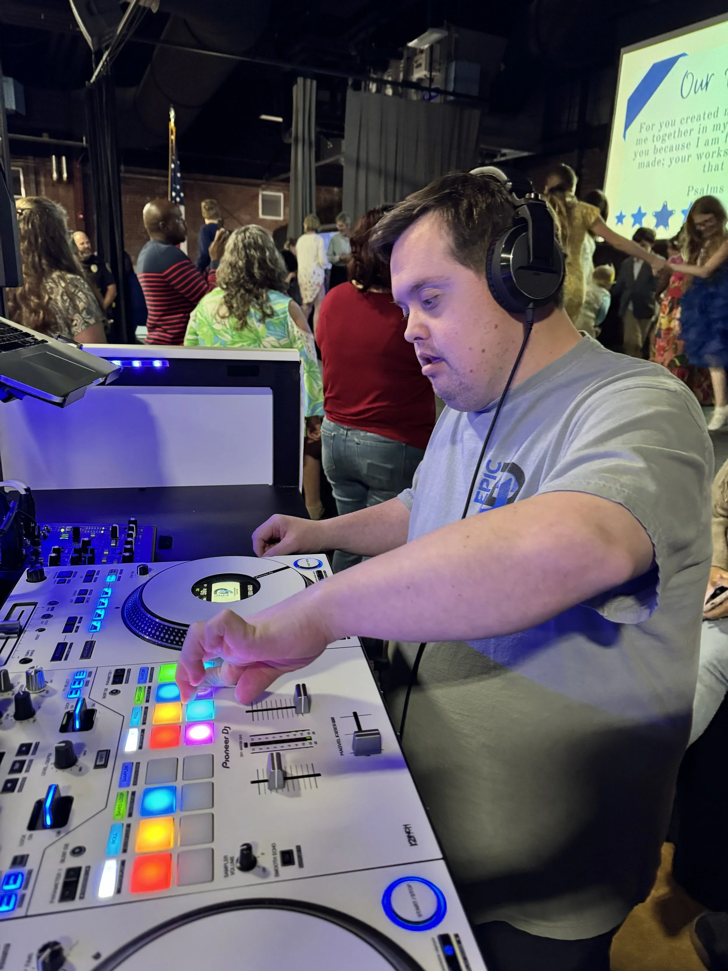 DJ mixing music at an event with a crowd in the background, large screen with text, and the DJ wearing headphones.