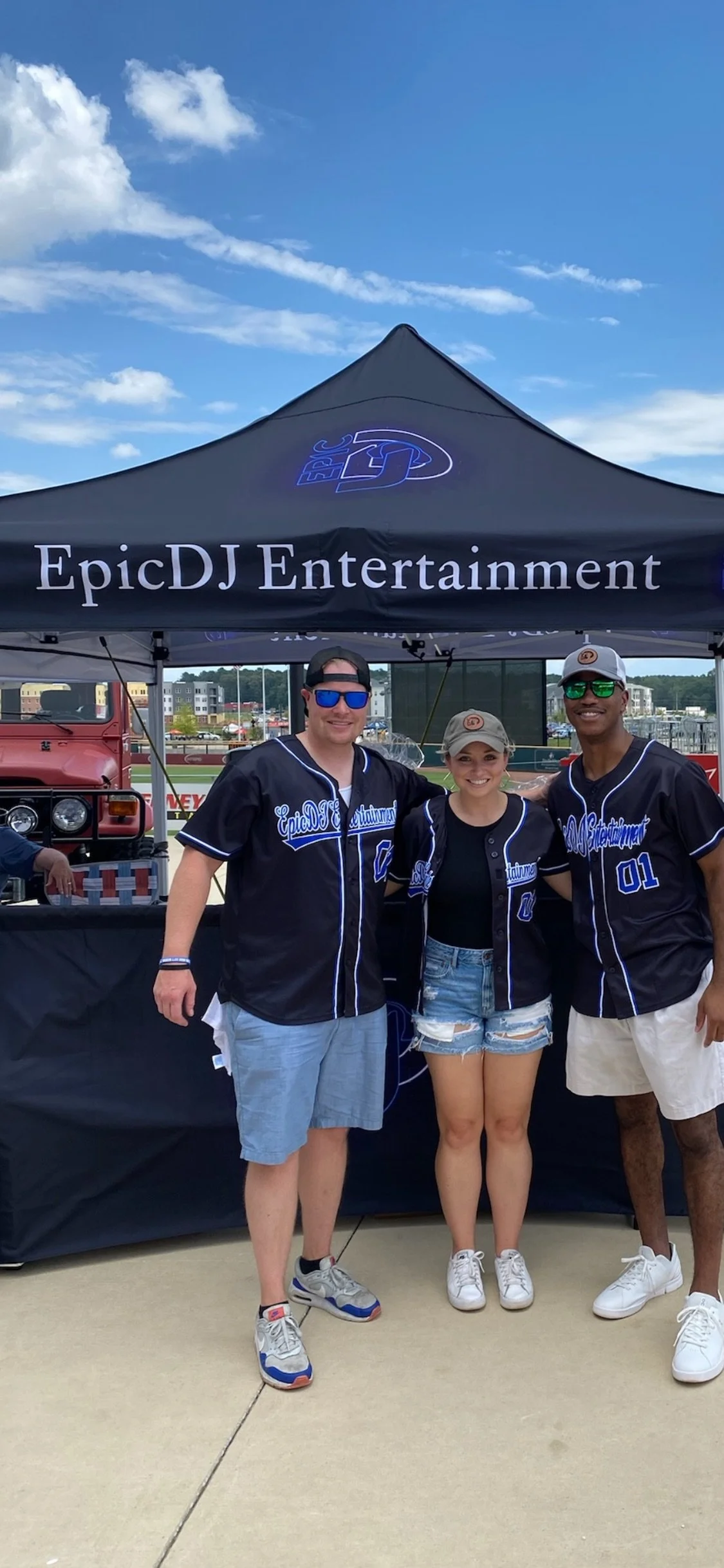 Three people in baseball jerseys standing in front of a tent with 'EpicDJ Entertainment' written on it at an outdoor event.