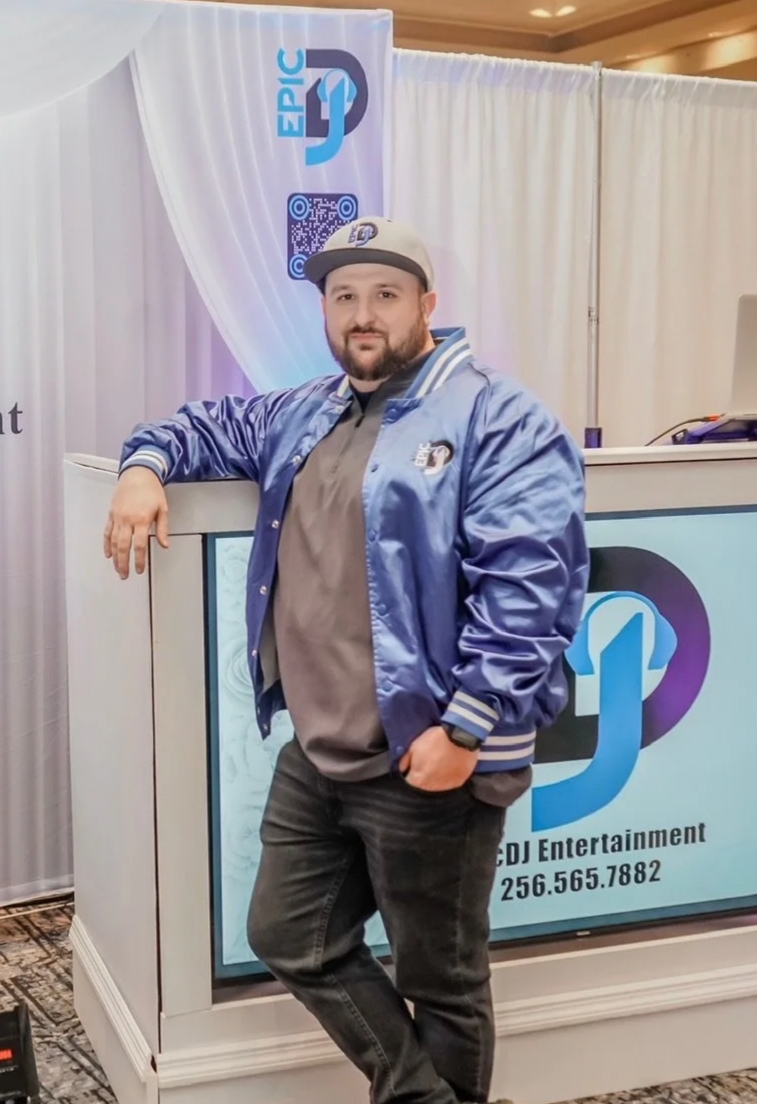 A man with a beard and mustache wearing a baseball cap, standing next to a DJ booth with a Carlton logo, in front of a banner that reads 'EPIC DJ' and 'DJ Entertainment' with a contact number. He is dressed in a gray t-shirt and a blue satin bomber jacket, with one hand resting on the DJ booth and the other in his pocket.