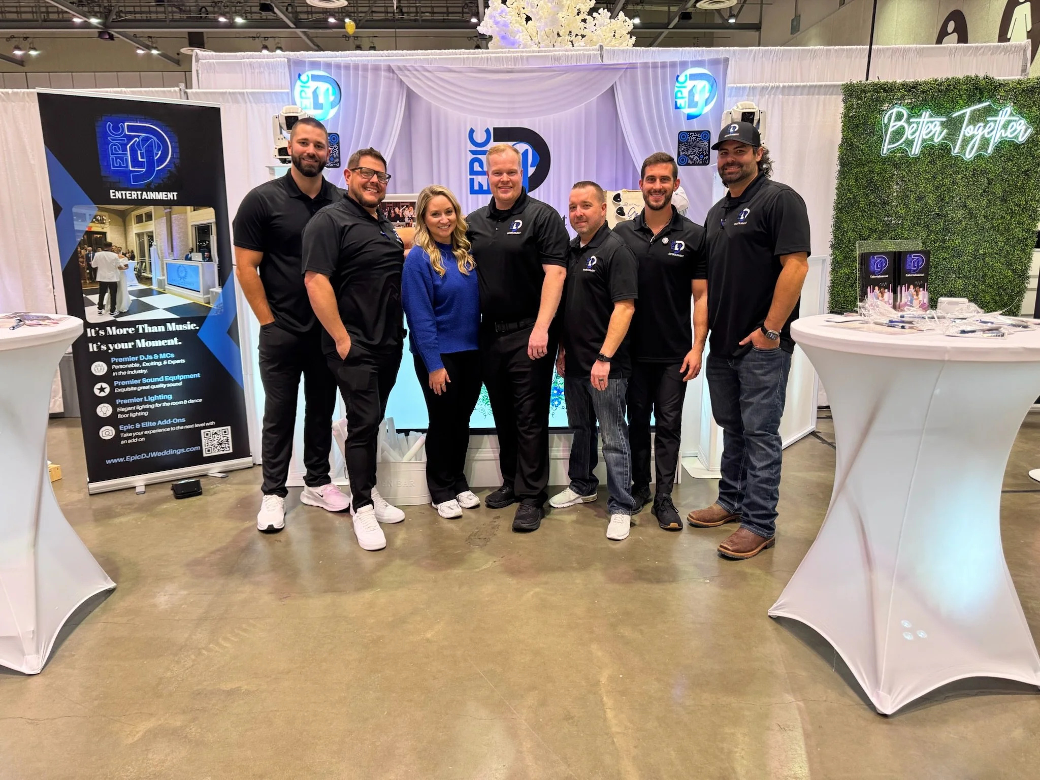 Group of eight people standing together in front of an Epic DJ booth at a trade show or event, with promotional banners and tables around them.