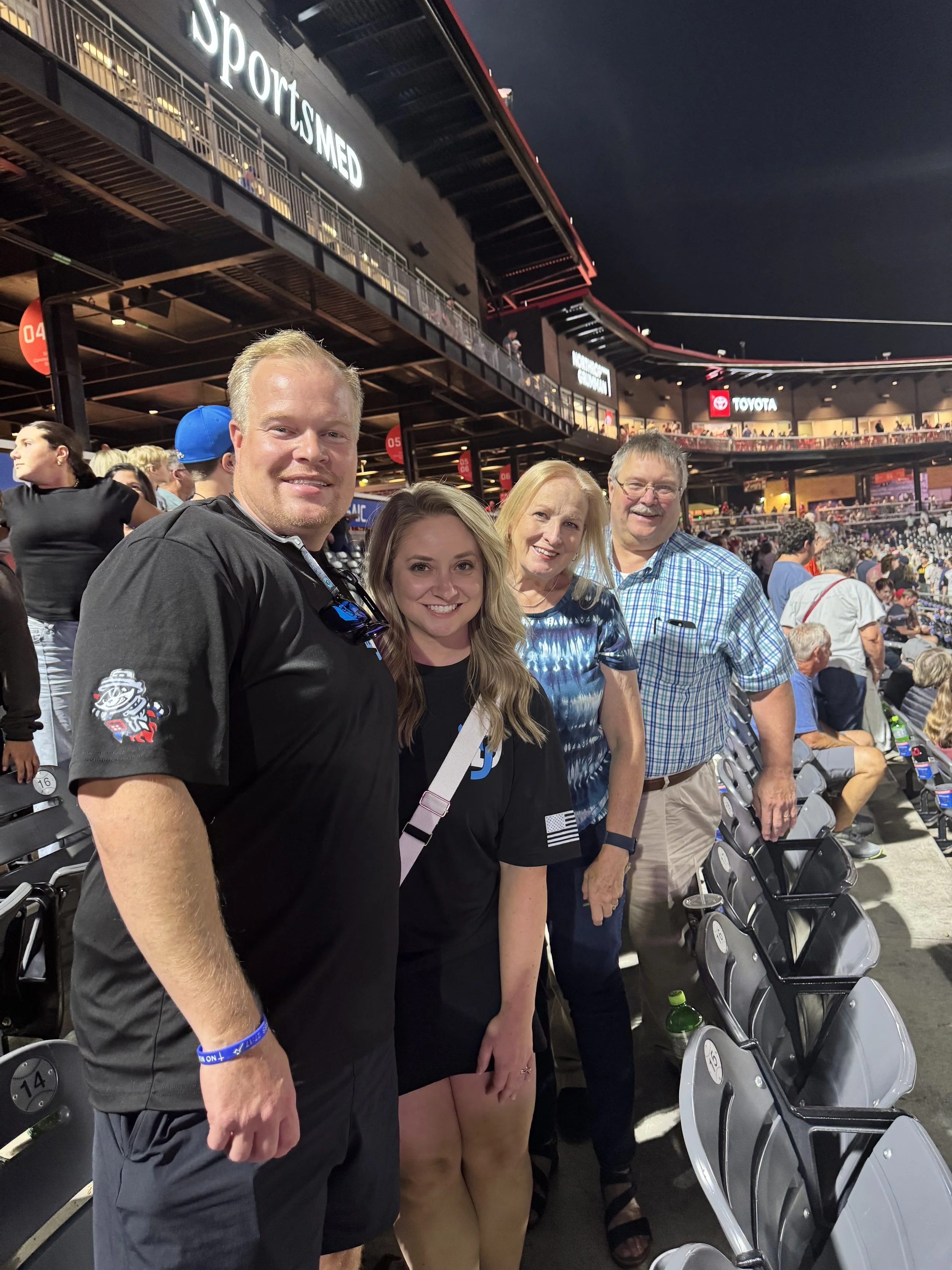 Four people standing together at a sports stadium during a baseball game, smiling, in the evening. The stadium is filled with other spectators and has large advertisements visible in the background.