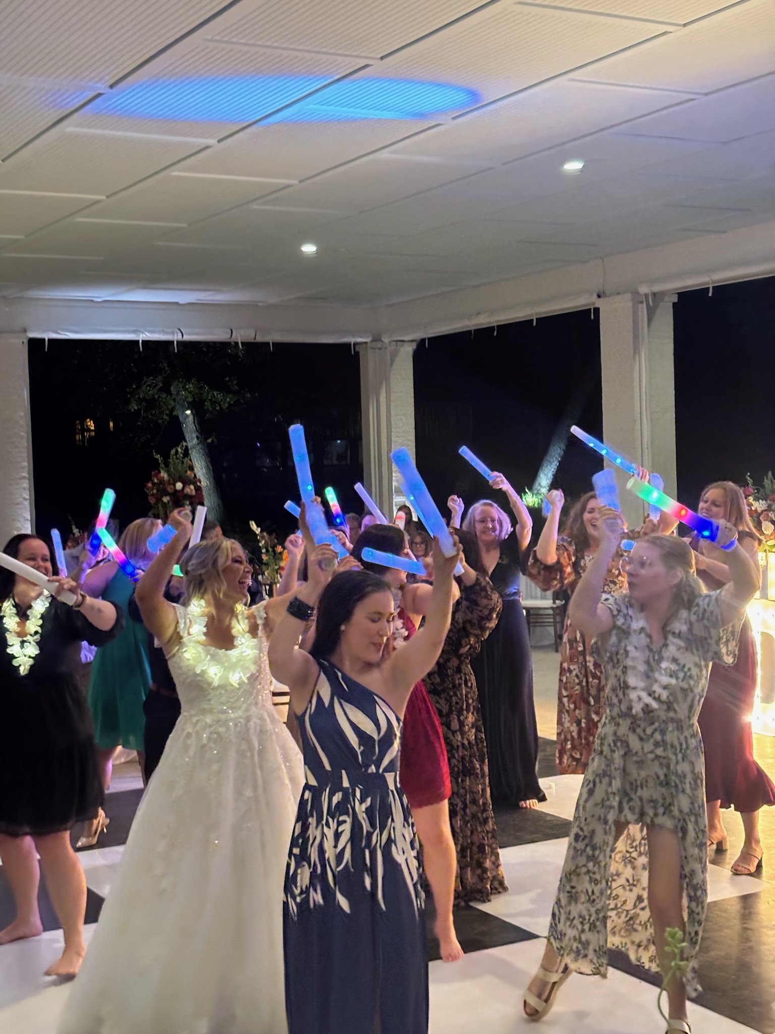 Group of women dancing at a celebration, holding colorful glow sticks, in a decorated indoor venue.