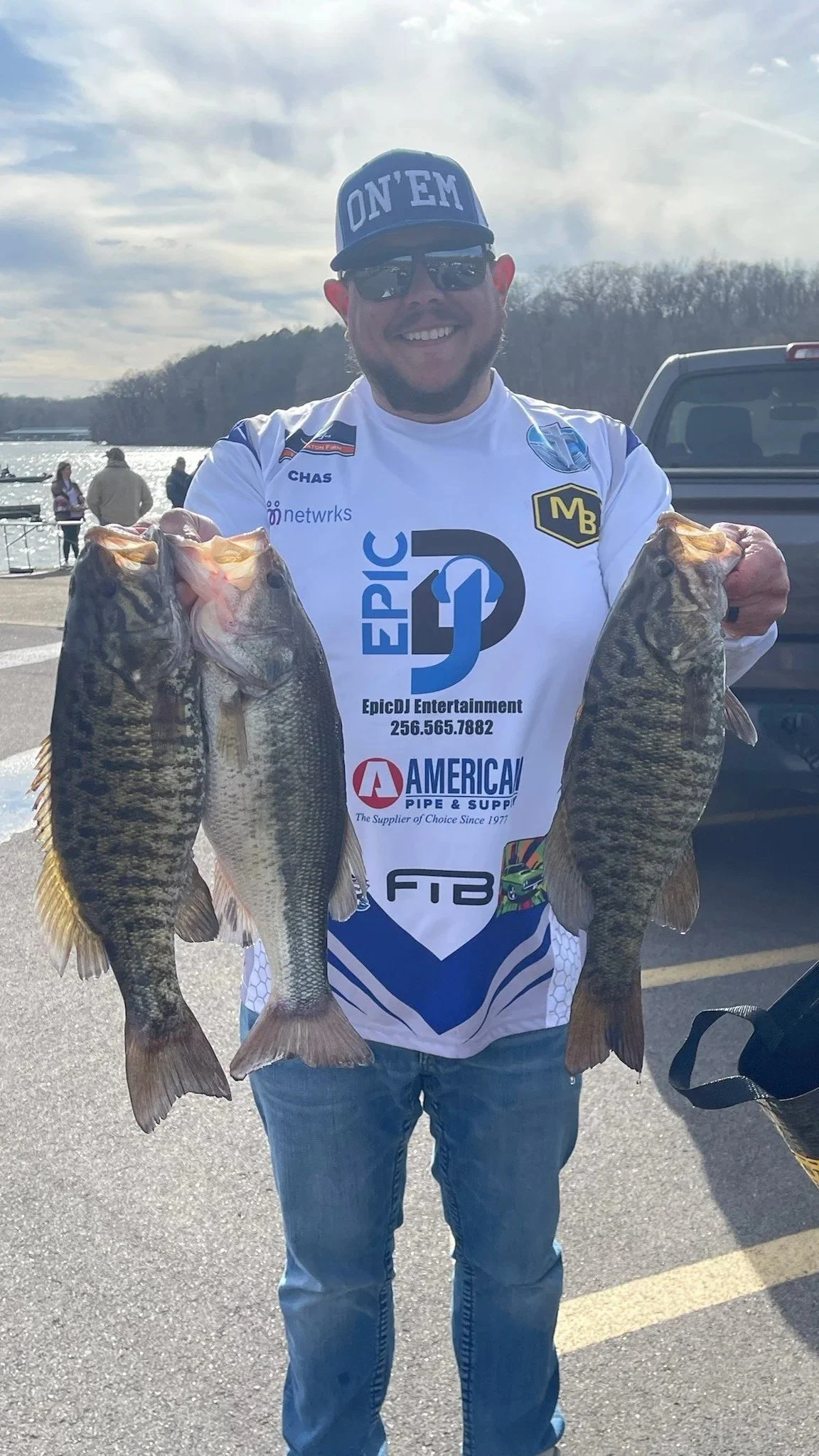 A man smiling and wearing a white fishing jersey, holding two large fish, standing outdoors near a body of water with other people and a boat in the background on a sunny day.