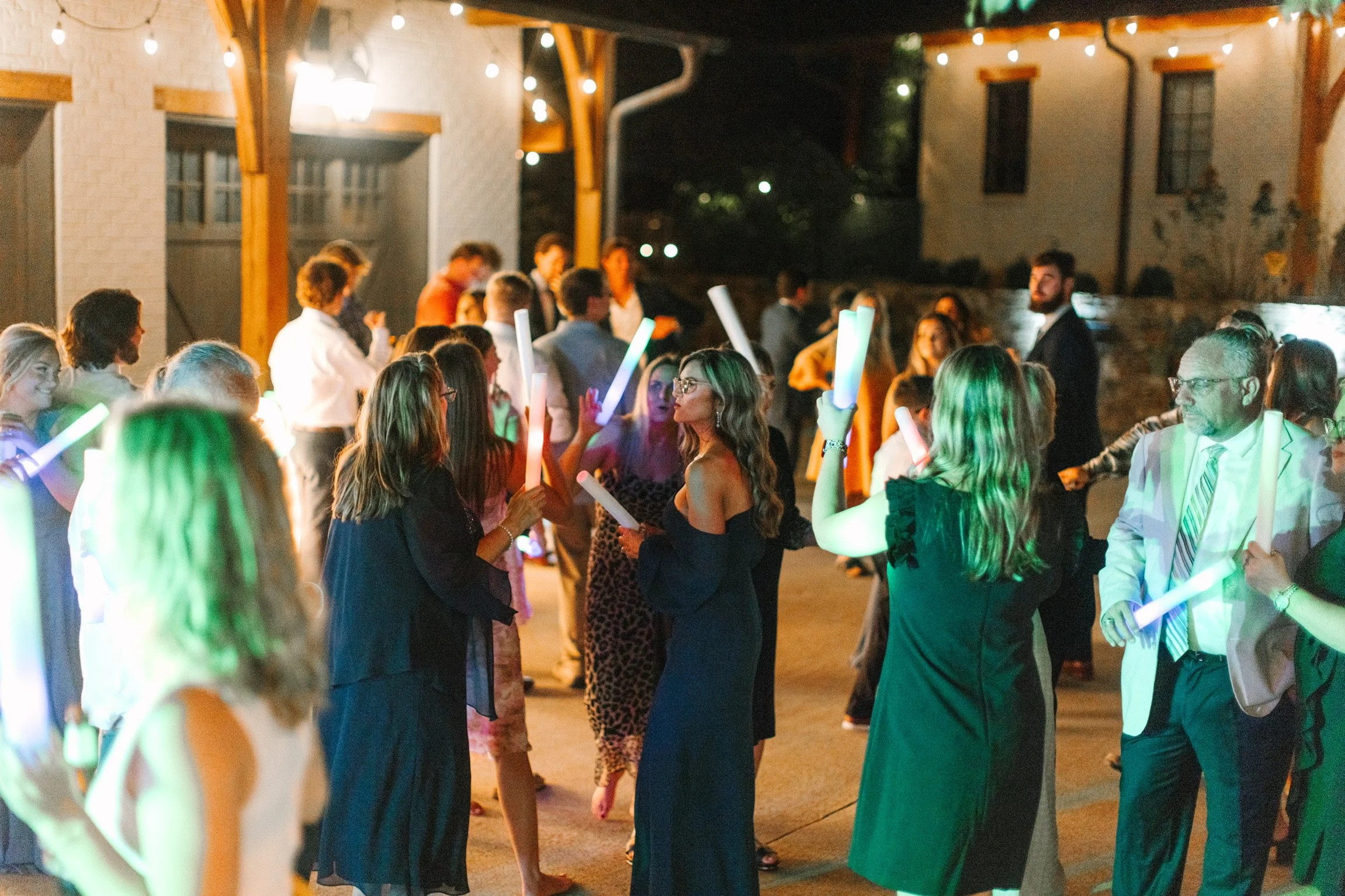 People dancing at an outdoor party with string lights, holding glow sticks, in a courtyard with wooden beams and brick walls at night.