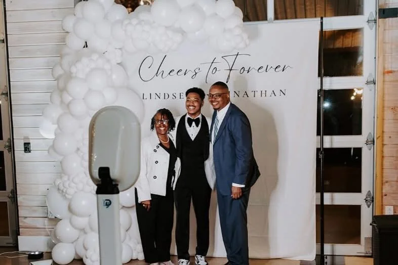 Three people standing together, smiling in front of a backdrop with balloons and the text "Cheers to Forever" and names Lindsay and Nathan, at a celebration or wedding event.
