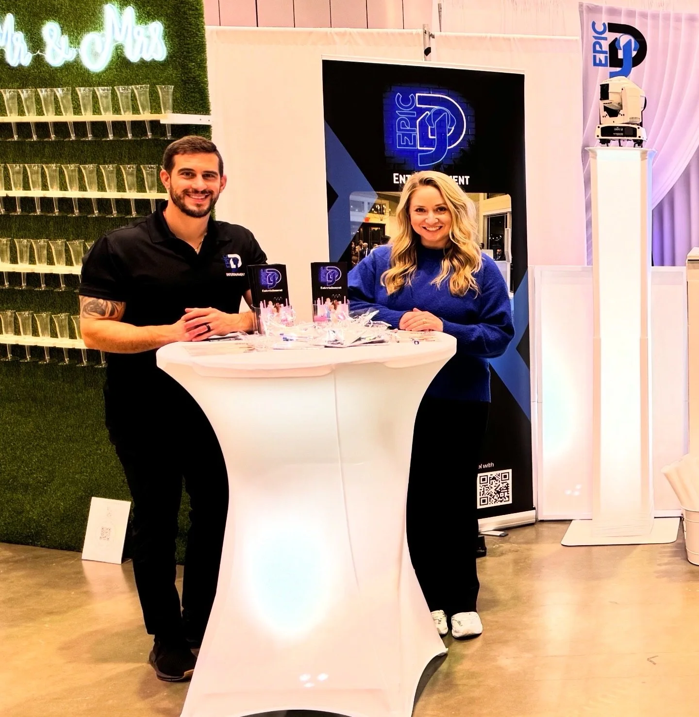 A man and woman standing behind a tall white table at an event booth for Epic DJ Entertainment, smiling at the camera.