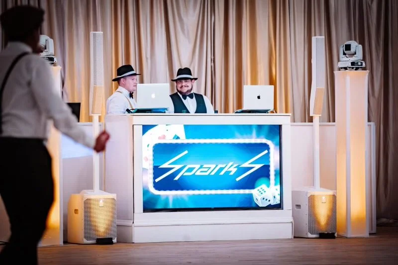 Two men wearing black hats and bow ties sitting behind a DJ booth with a large screen displaying 'Spark' and two laptops. A woman in the foreground is barely visible.
