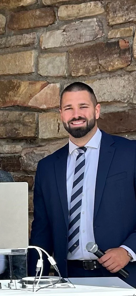 A man with a beard dressed in a dark blue suit, white shirt, and striped tie holding a microphone stands in front of a stone wall.
