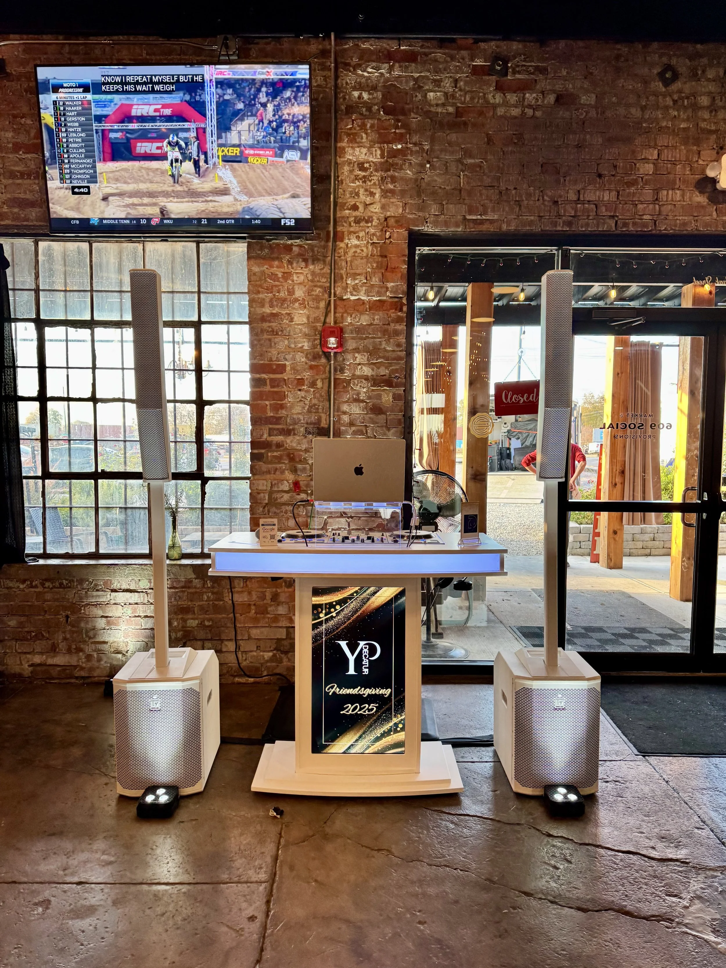 Interior of a venue with a brick wall, a DJ setup on a white table, large speakers on either side.