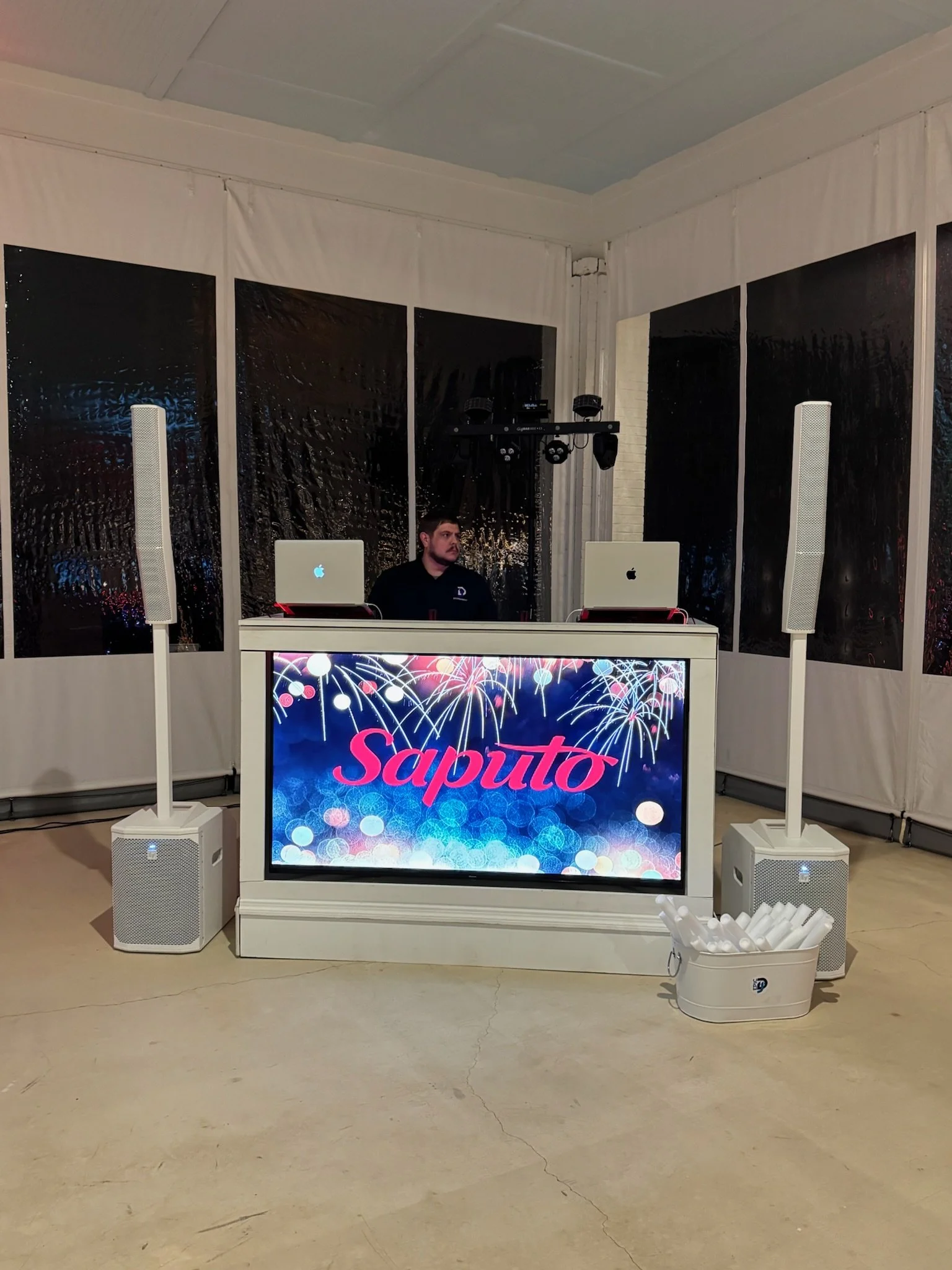 DJ booth with two laptops, large speakers, and a screen displaying 'Saputo' with fireworks background, set up indoors with black wall panels and a DJ person in the background.