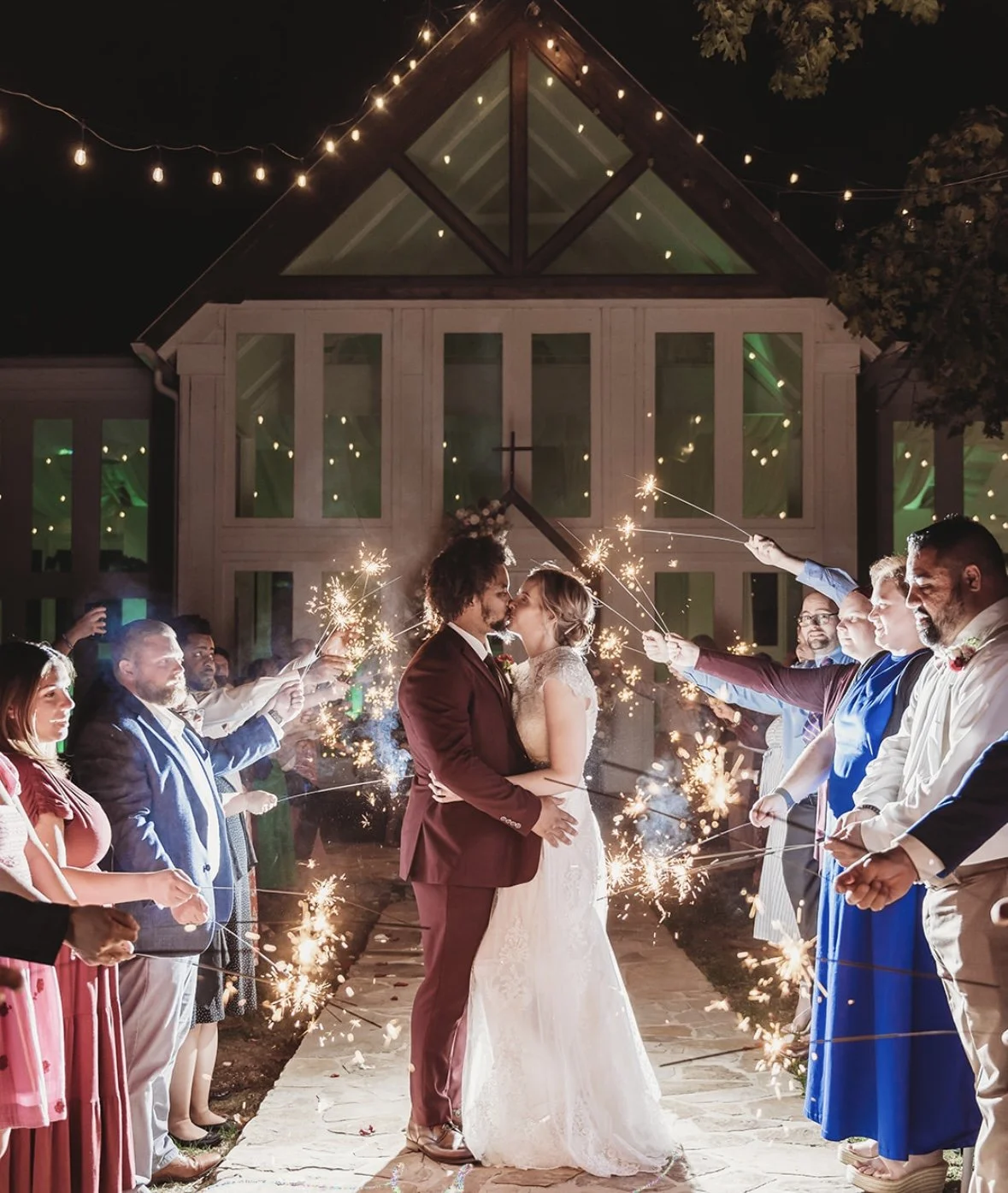 A bride and groom share a kiss during their wedding celebration, surrounded by guests holding sparklers at night, outside a lit building with string lights.