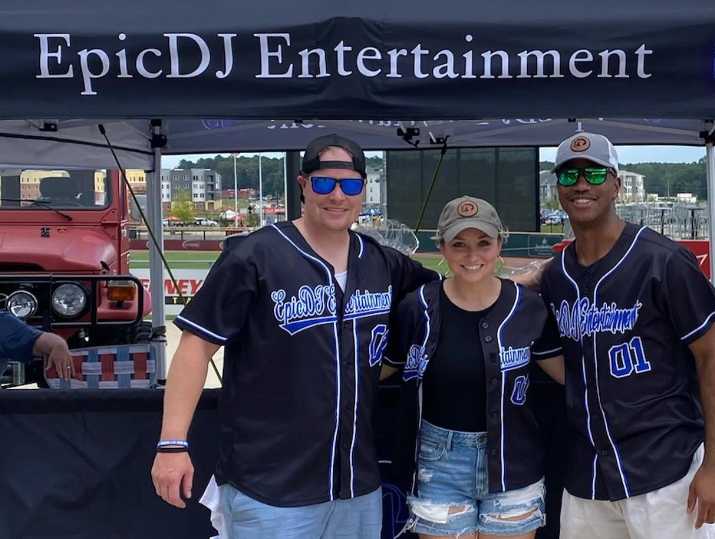 Three people wearing black EpIcDJ Entertainment jerseys with blue accents, posing under a black tent with the company's name, at an outdoor event near a baseball field, smiling at the camera.