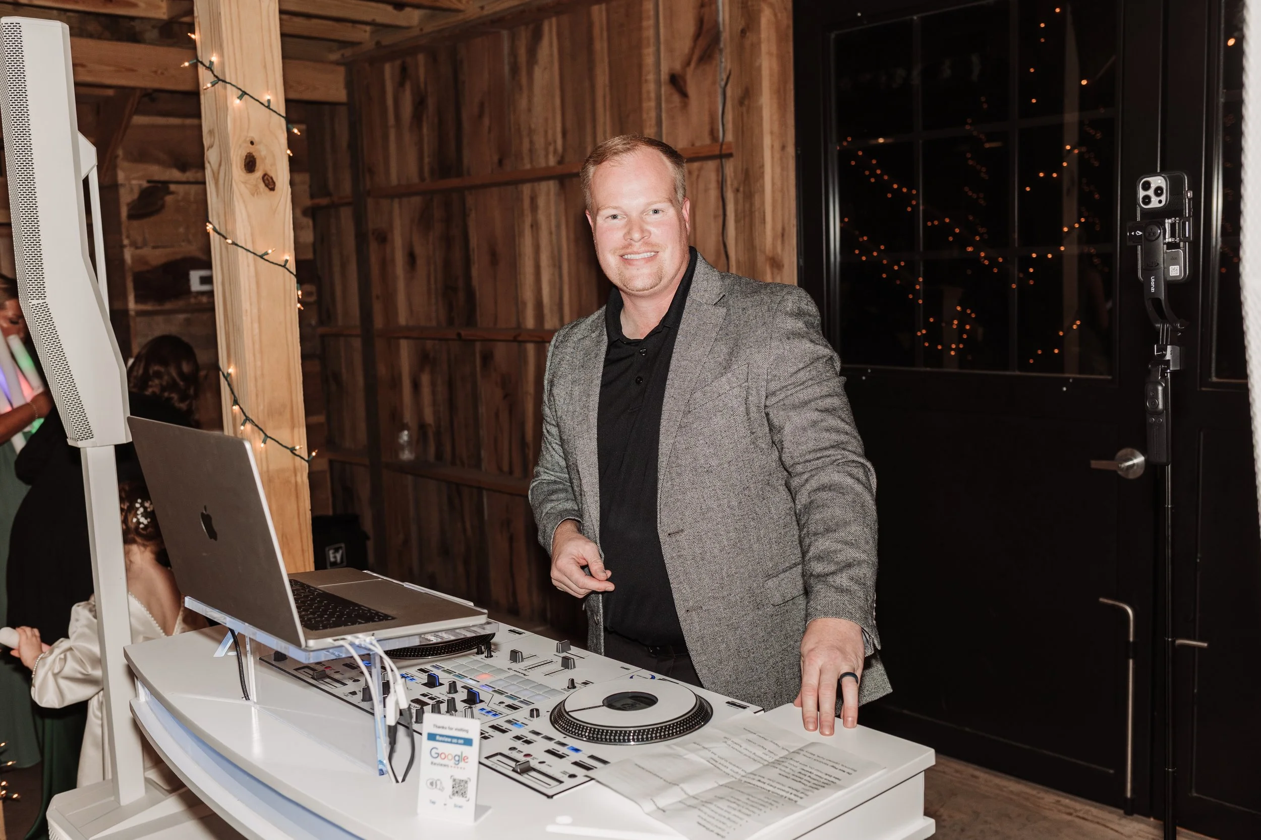A man smiling at a DJ setup with a laptop and music mixer, standing inside a rustic wooden event space decorated with string lights.