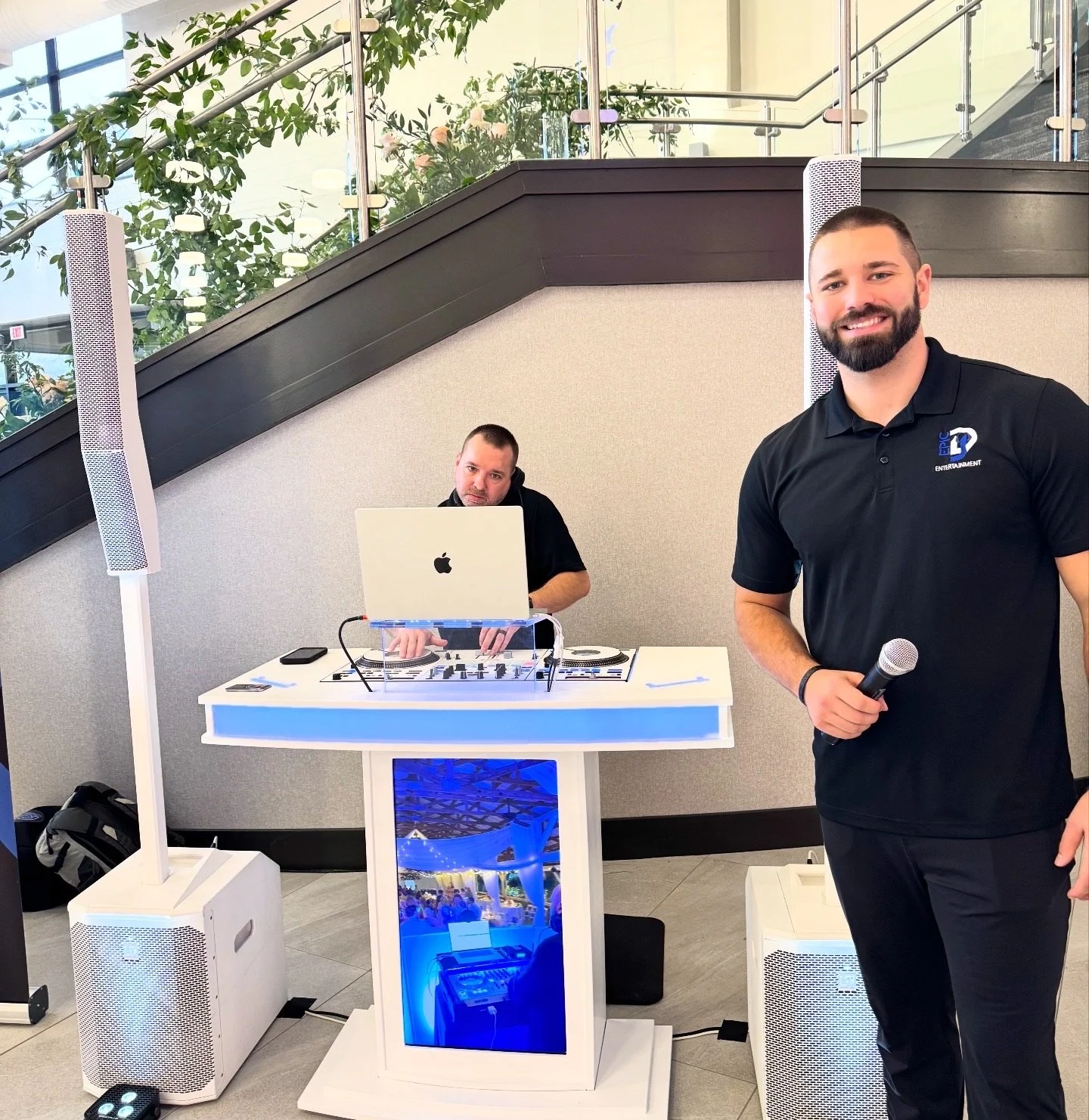 A man smiling and holding a microphone, standing in front of a DJ setup with another man working on a laptop and DJ equipment. The scene is indoors with plants and glass stairs in the background.