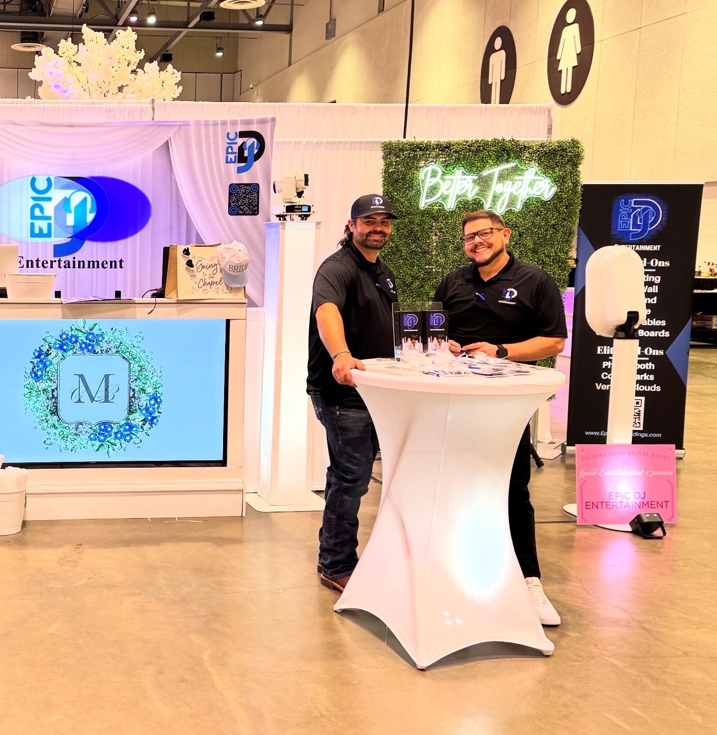 Two men standing at a round high-top table, smiling at the camera, at an exhibition booth with signs that read 'Epic DJ Entertainment' and 'Better Together'. 