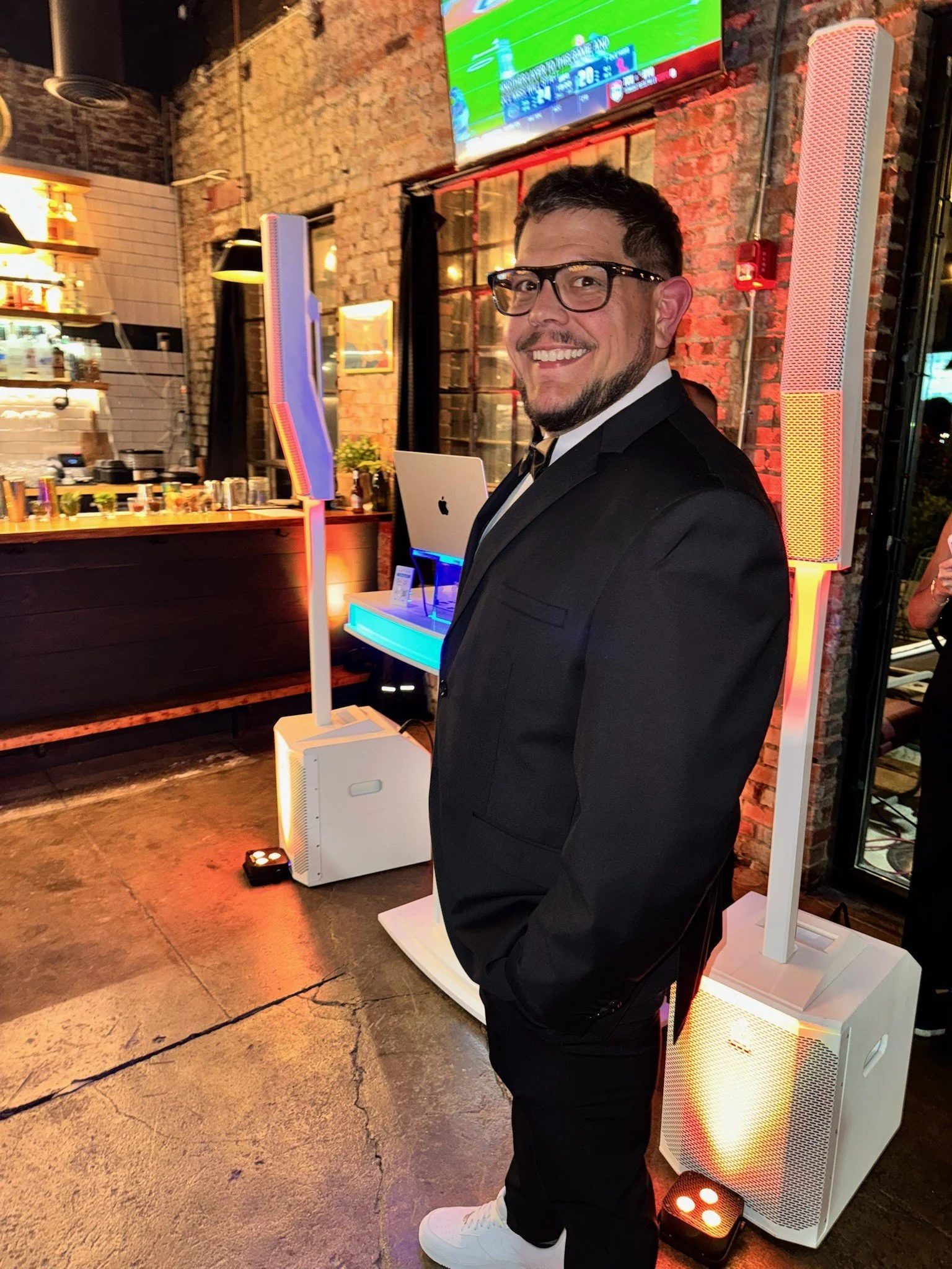 A man in a black tuxedo with glasses smiling at the camera at a social event in a brick-walled venue with a bar and a large screen television overhead.