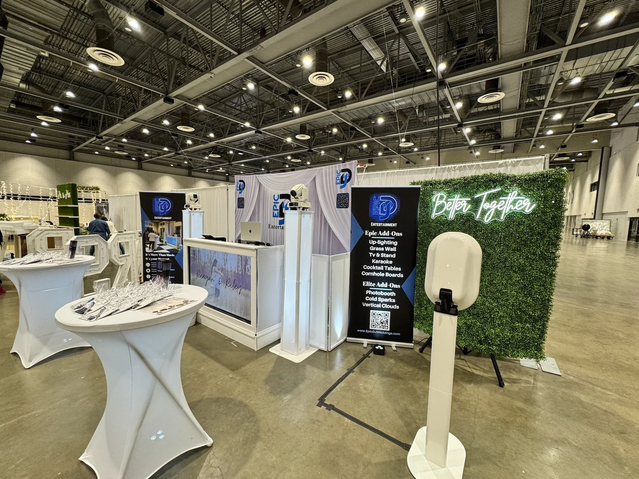 An exhibition booth at a trade show with a green hedge backdrop and neon sign that reads 'Better Together'. There are tables with glassware and promotional materials, a photo booth with scan code, and display banners for Epic Entertainment and their 