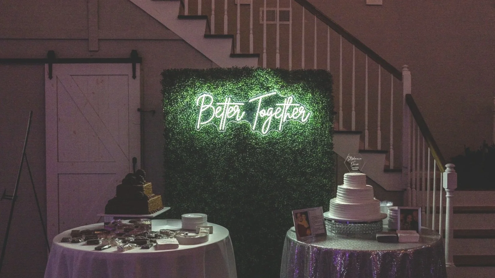Celebration setup with a neon sign that says 'Better Together' on a green leafy wall. There are two tables with desserts: one with a multi-layered white cake and framed photos, and another with a chocolate cake and various treats. The scene is indoors near a staircase.