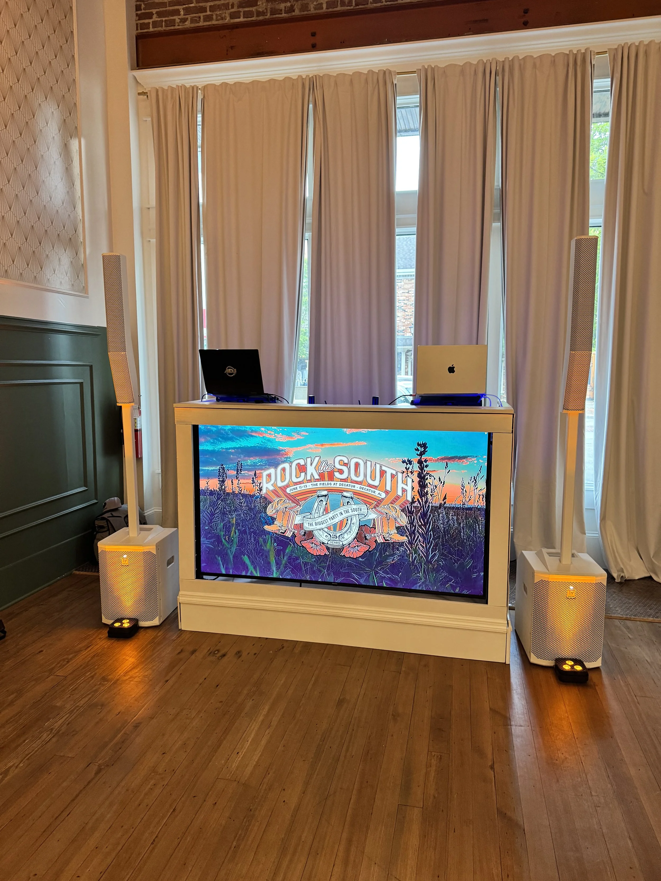 A DJ setup with a large screen displaying a colorful promotional graphic for 'Rock the South'. Two laptops are on top of the white DJ booth, with speakers on either side placed on the floor. The room has hardwood floors, white curtains, and windows i