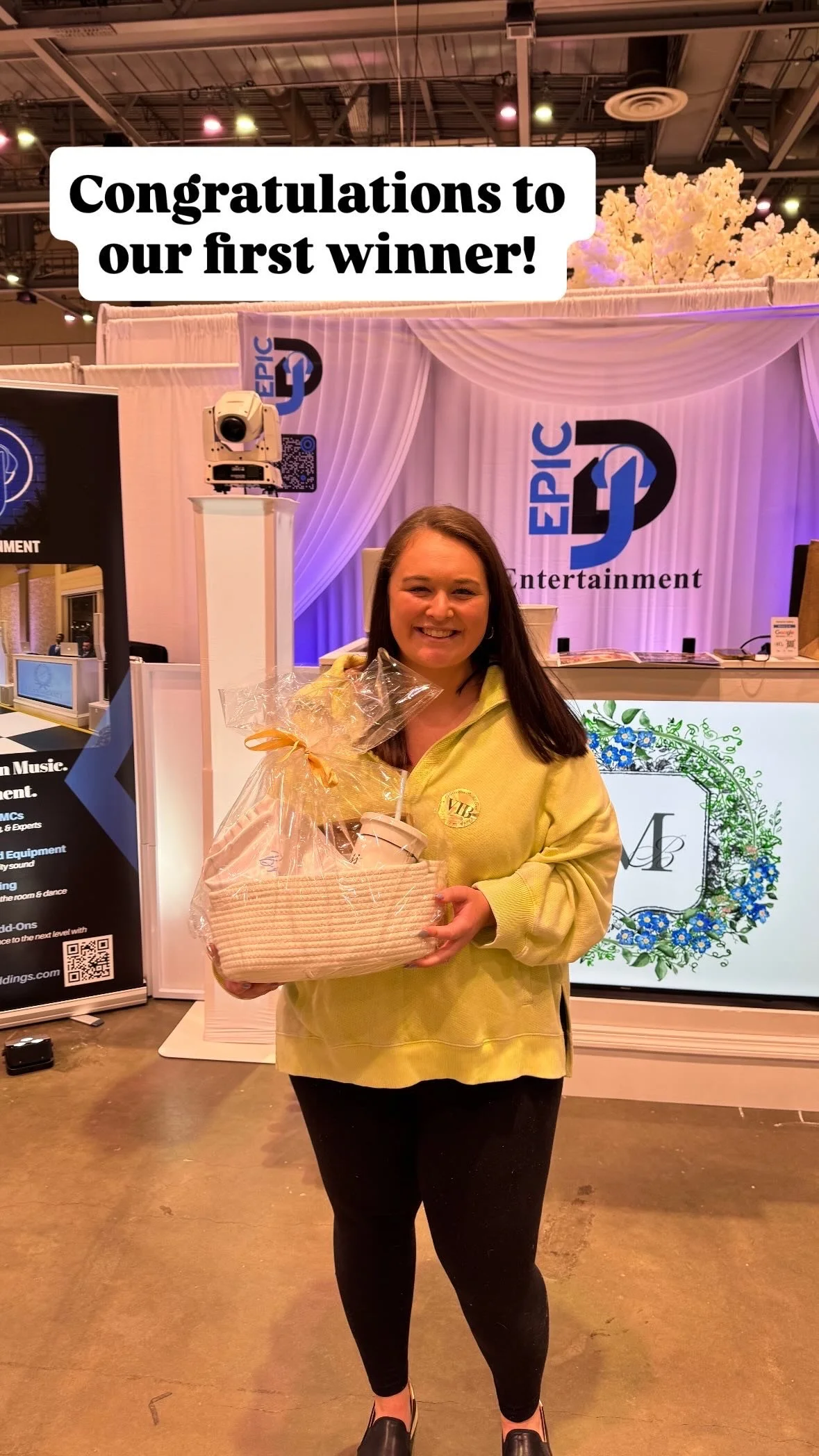 A woman smiling and holding a gift basket with wrapped items inside at an event, with a backdrop featuring the EPIC Entertainment logo and decorative drapery.
