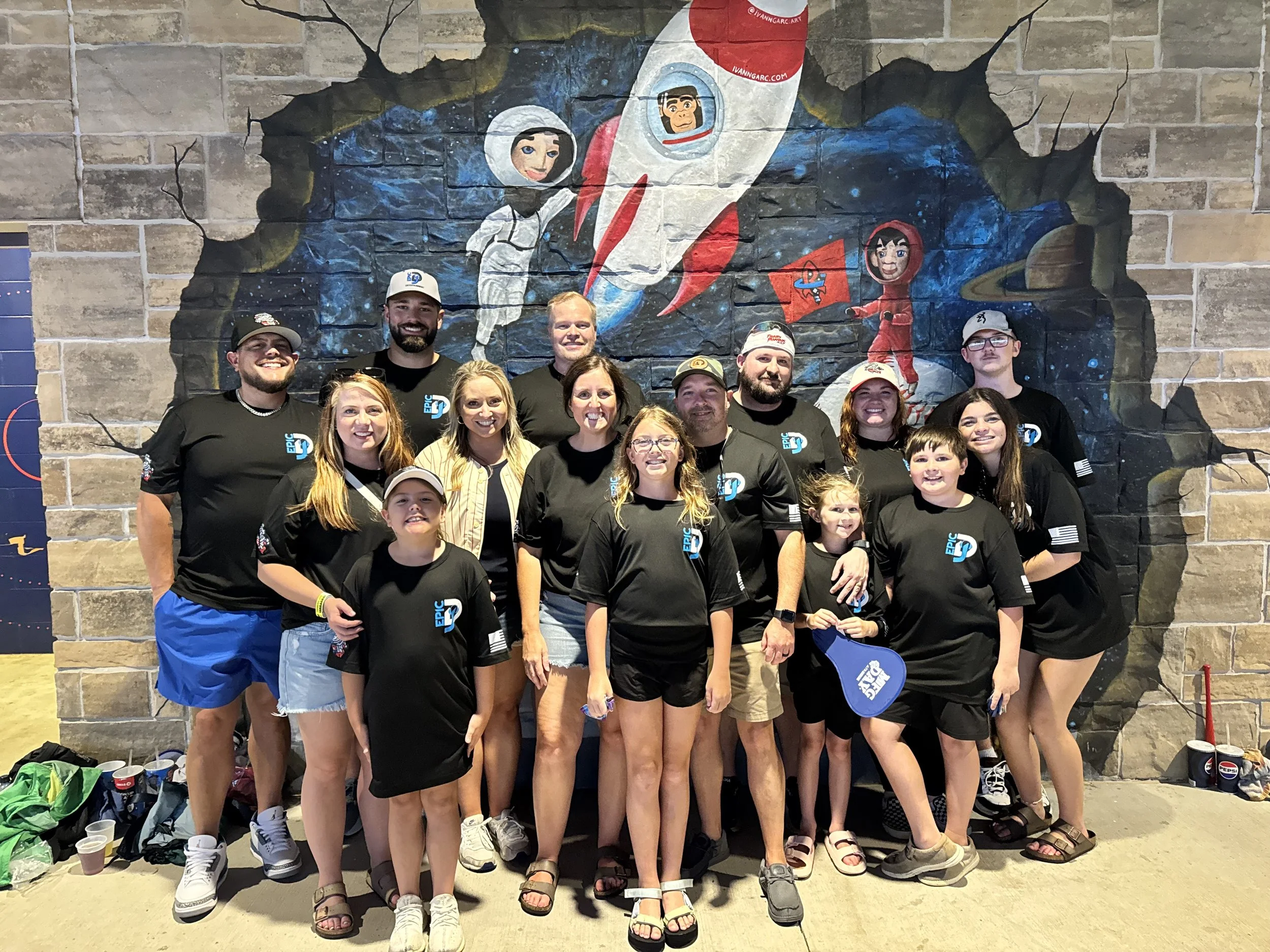 A group of people, including children and adults, posing in front of a mural with space and rocket themes at an indoor venue. Most are wearing black shirts with a blue and white logo.