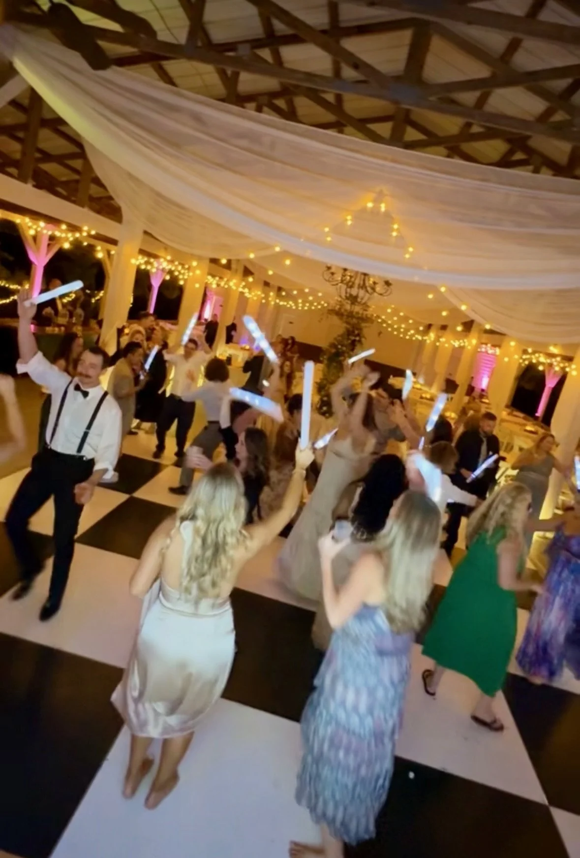 People dancing at a wedding reception or party with string lights and draped fabric ceiling decorations, on a black-and-white checkered dance floor.