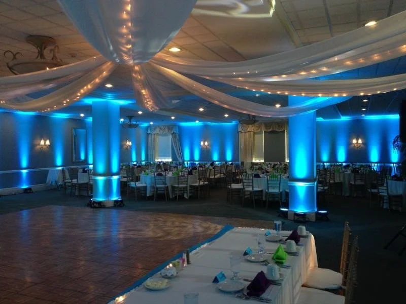 An elegant banquet hall decorated with white drapery and blue uplighting, featuring round tables and a dance floor.