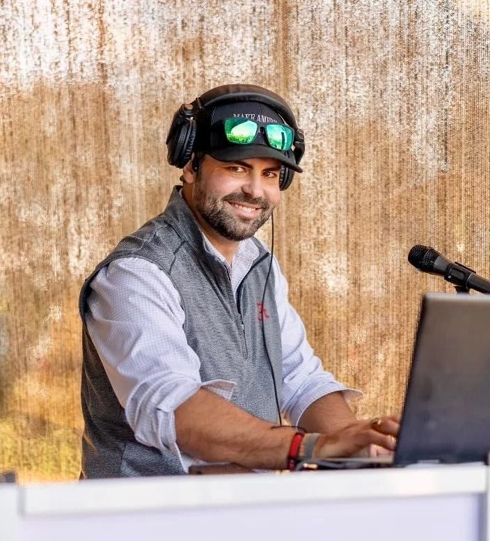 A man smiling while sitting at a table with a laptop, wearing headphones, a cap with sunglasses on top, and a light-colored shirt with rolled-up sleeves, in front of a textured background.