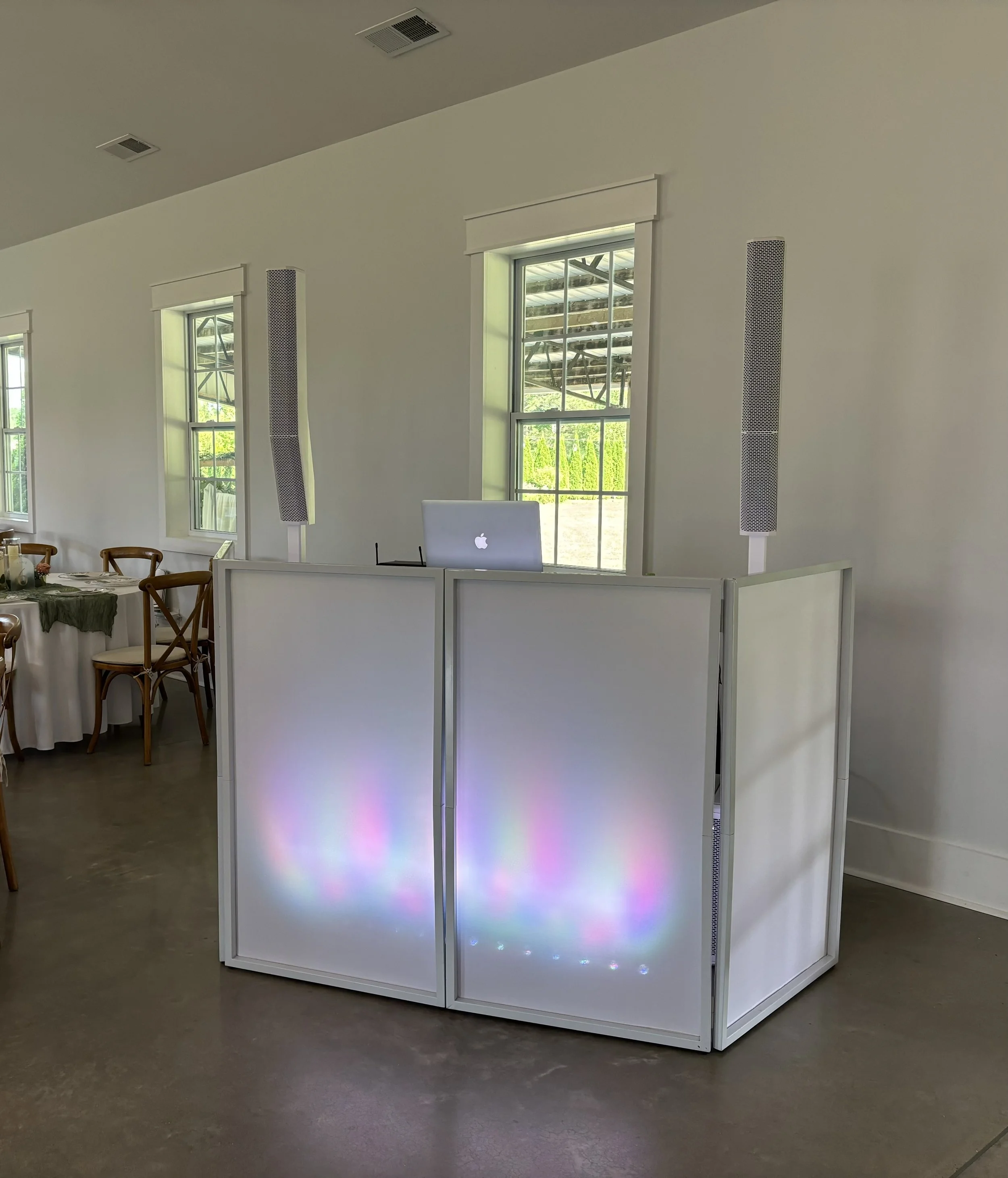 A DJ setup with a white foldable partition, a laptop on top, and two tall speakers in a bright room with windows.