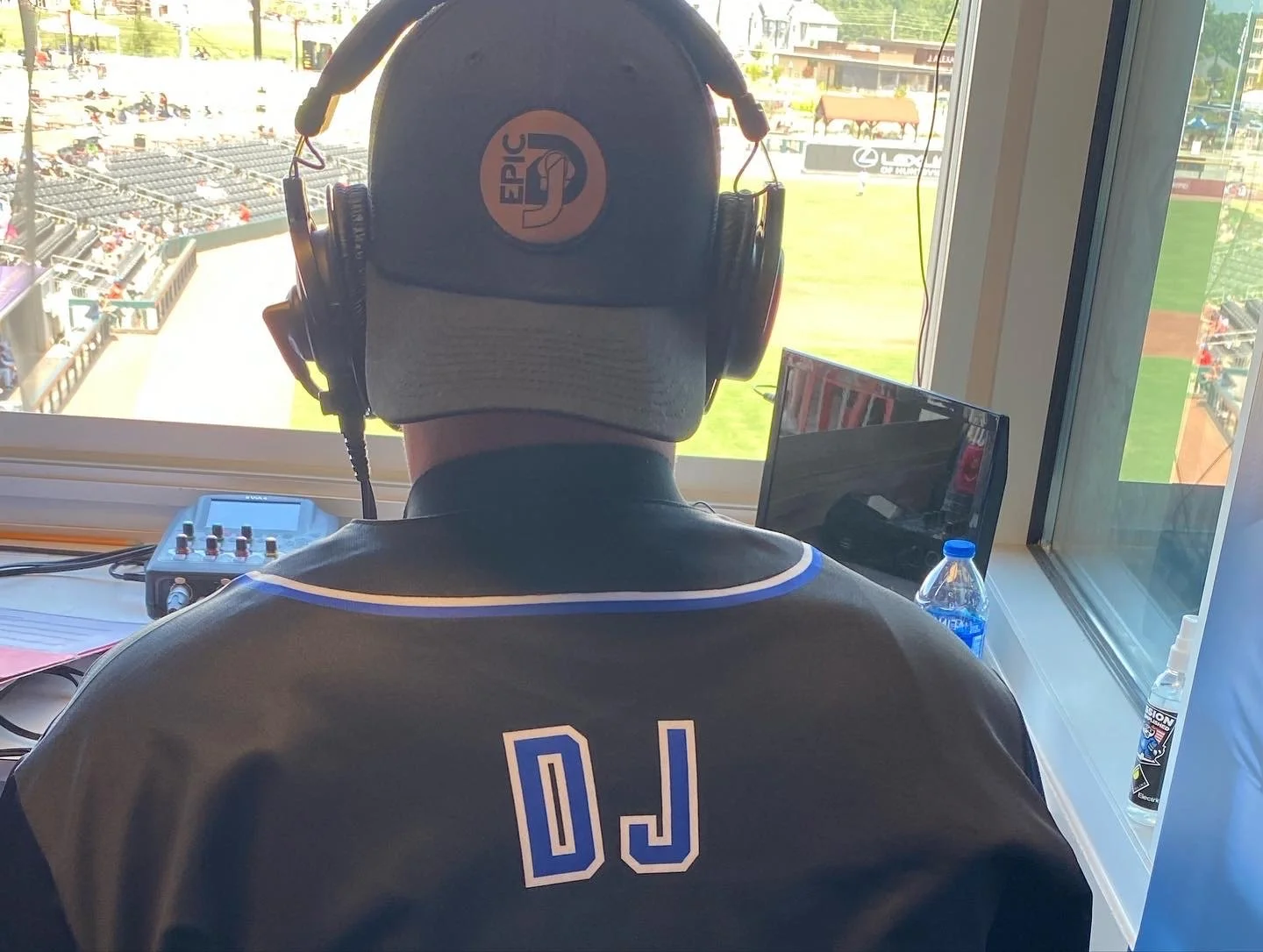 Back view of a DJ wearing a black jacket with 'DJ' printed on the back, DJ wearing headphones with 'Epic' logo, operating DJ equipment near a window overlooking a sports stadium, water bottle and electronic device on the table.