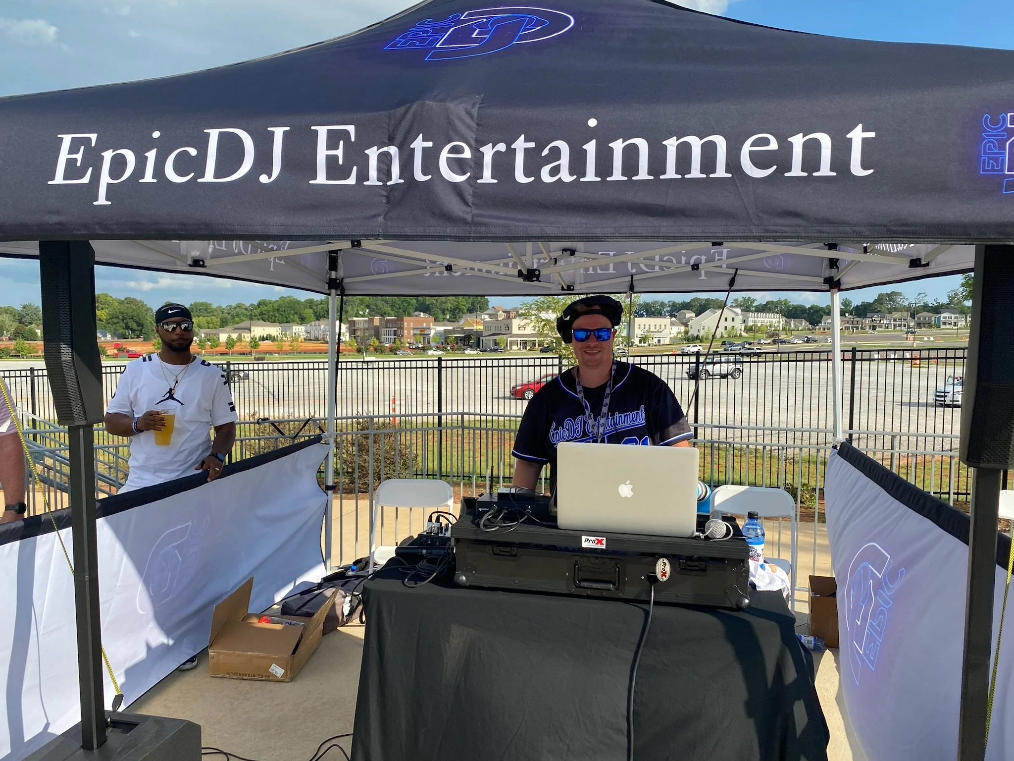 A DJ booth with two people in a tent, one DJ wearing sunglasses and a black shirt, the other with a white shirt and sunglasses, outdoors with a parking lot and residential buildings in the background.