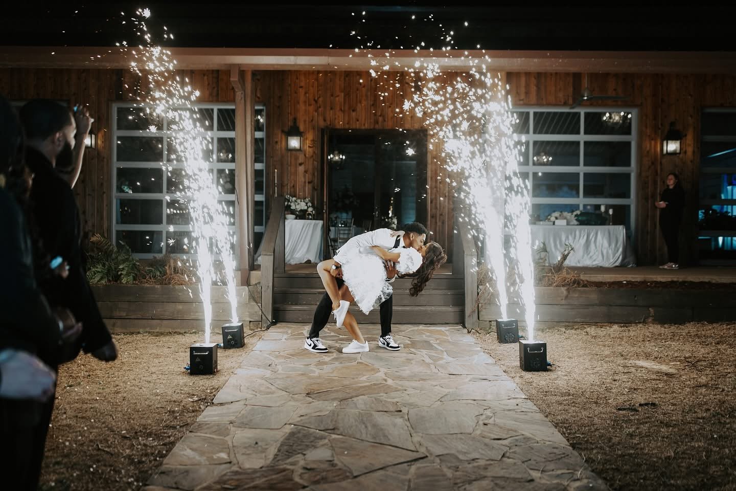 Couple dancing with sparks flying on either side during a celebration outside a wooden building at night.