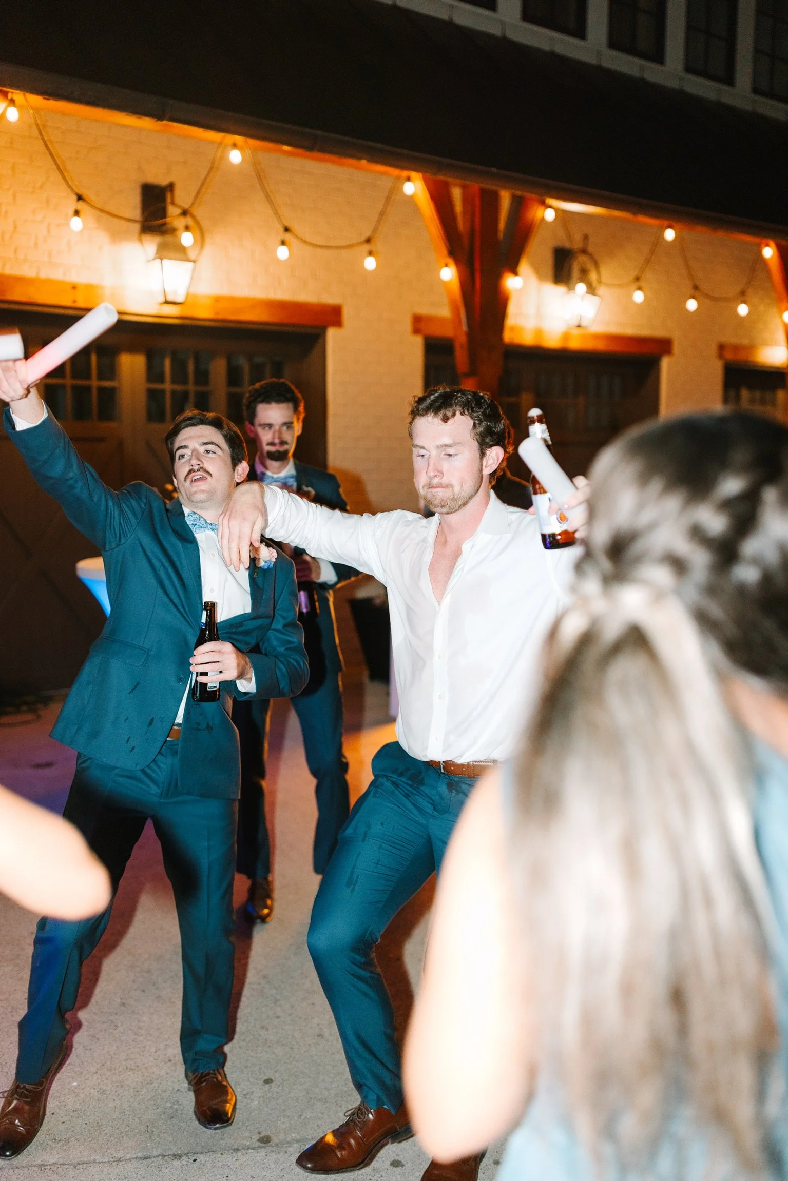People dancing and having fun at an outdoor party or celebration at night with string lights overhead, some holding drinks.