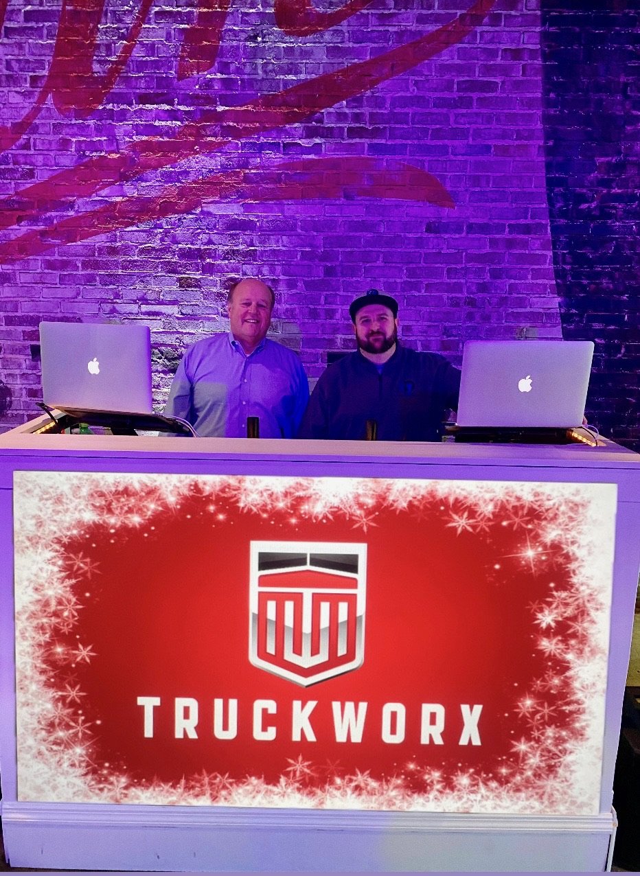 Two men standing behind a DJ booth with Apple MacBook laptops, a brick wall with purple lighting, and a red sign with white snowflake decorations displaying TrucketWorx logo.