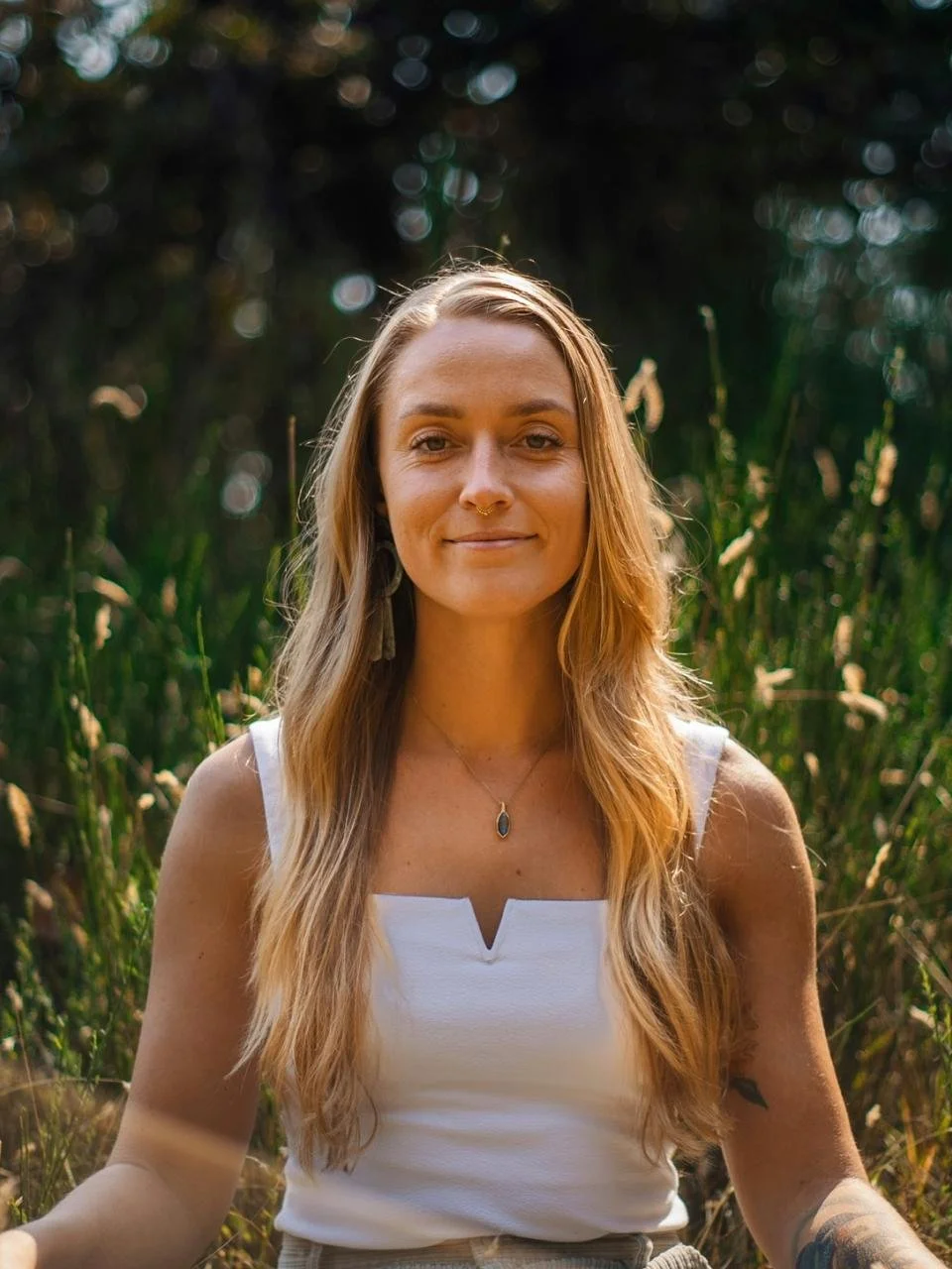 A woman with long, wavy blonde hair wearing a white sleeveless top and jewelry, smiling and sitting outdoors in a natural setting with greenery and blurred trees in the background.