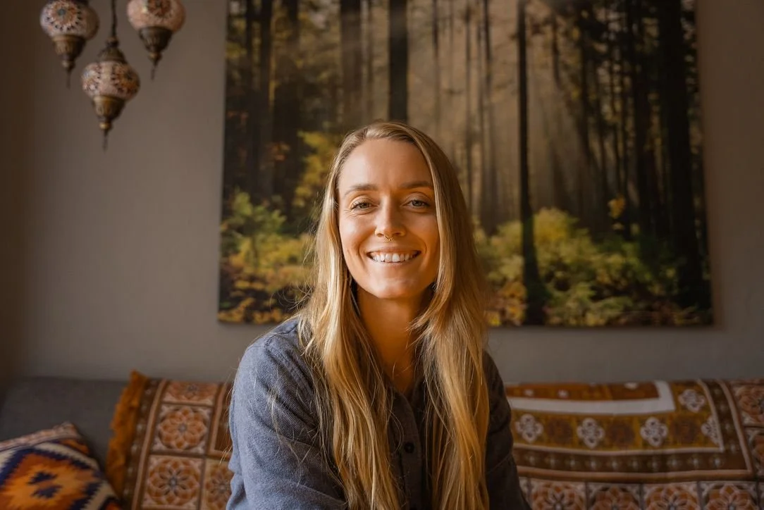 A smiling woman with long blonde hair sitting on a patterned sofa in a room with a forest scene tapestry in the background.