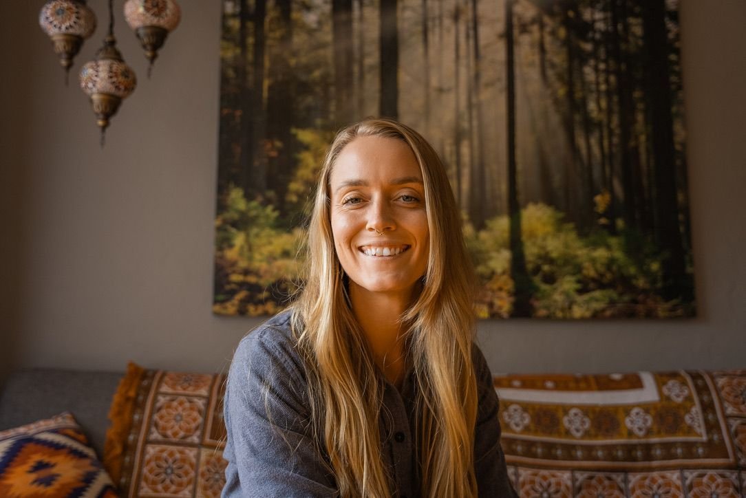 A woman with long blonde hair smiling indoors, sitting on a sofa with patterned cushions, background features a large forest landscape photo on the wall, and hanging decorative lamps.
