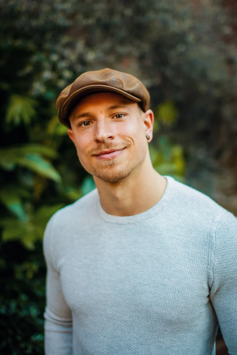 Portrait of a young man wearing a brown cap and a light gray long-sleeve shirt, standing outdoors with green foliage background.