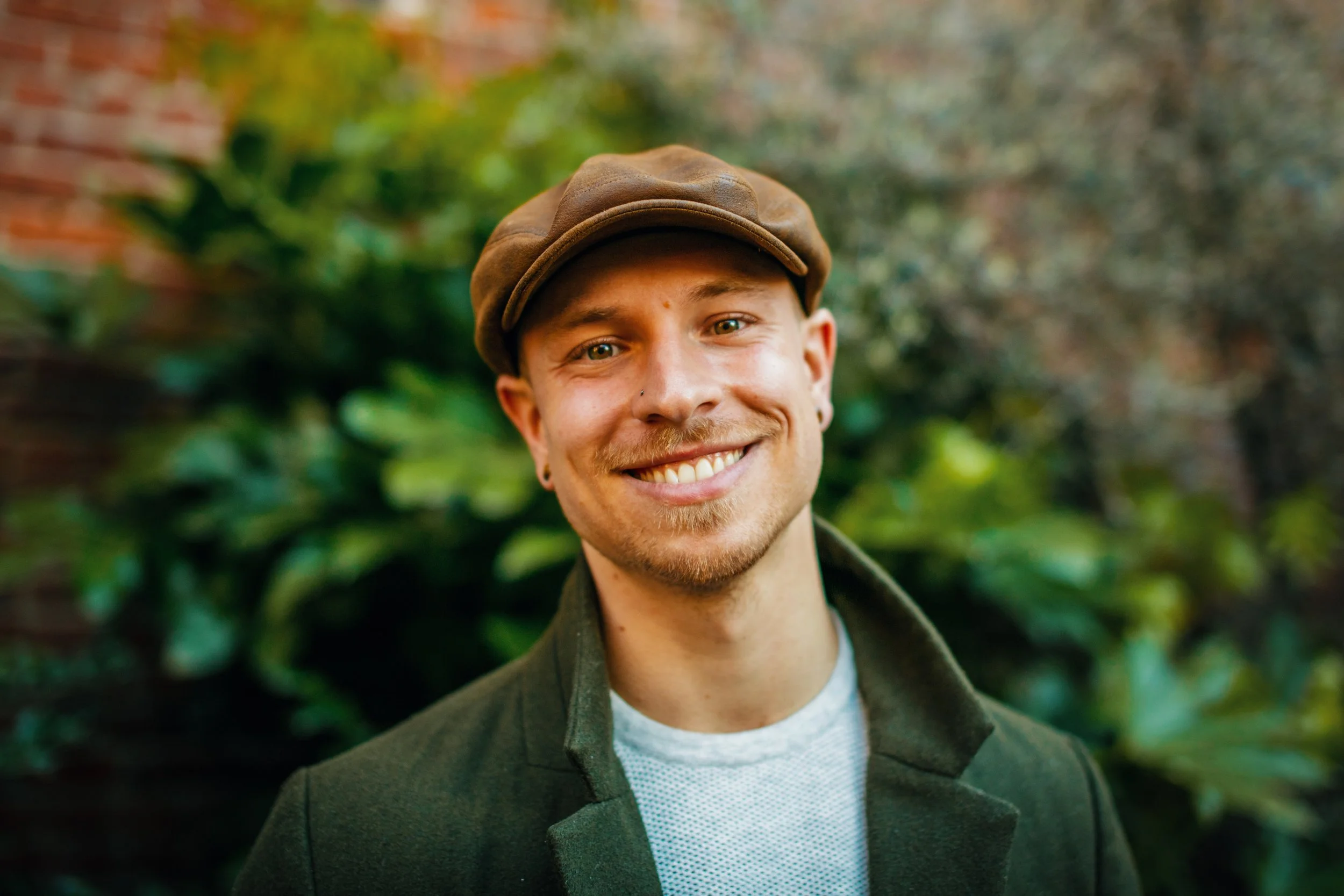 A smiling man with a beard and nose piercing, wearing a brown hat and dark coat, standing outdoors with green foliage in the background.