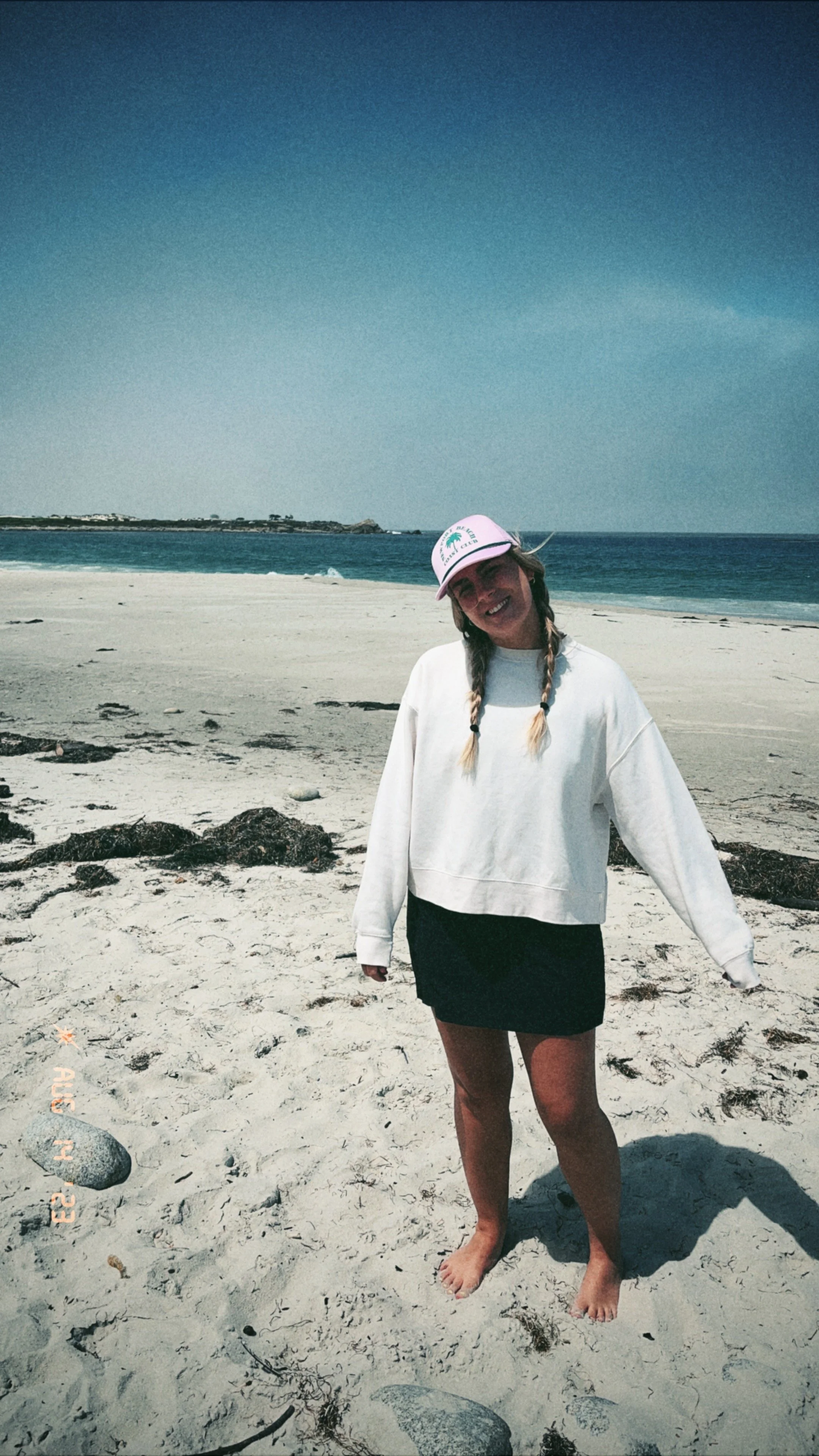 A person standing barefoot on a sandy beach, smiling, with ocean waves and a clear sky in the background. They are wearing a white sweatshirt, a black skirt, and a pink cap. Founder of Already Coolest Studio, Samantha.