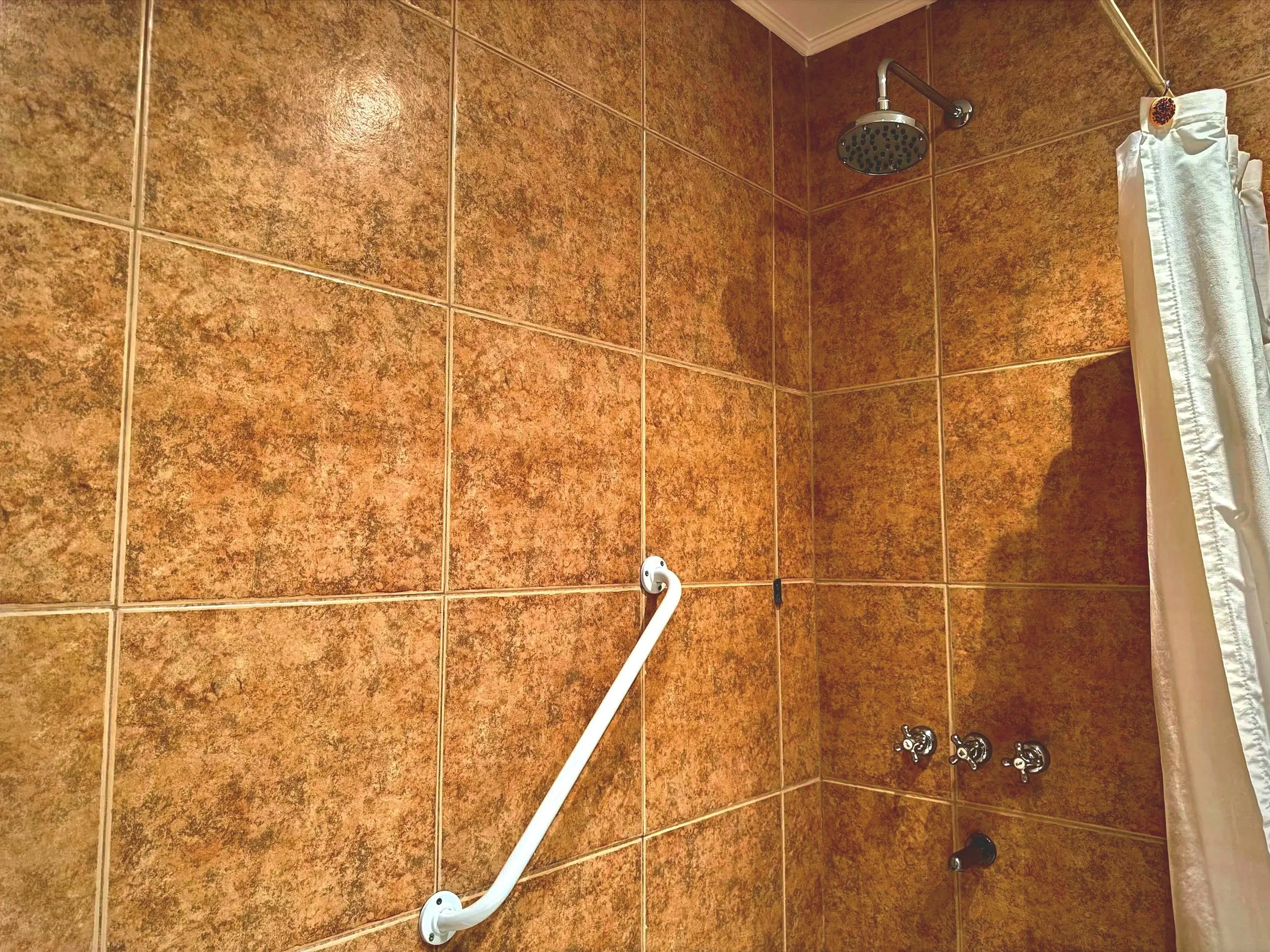 Vista de un baño con azulejos marrones en las paredes, una ducha con cabezal de lluvia y una cortina blanca en el lado derecho.