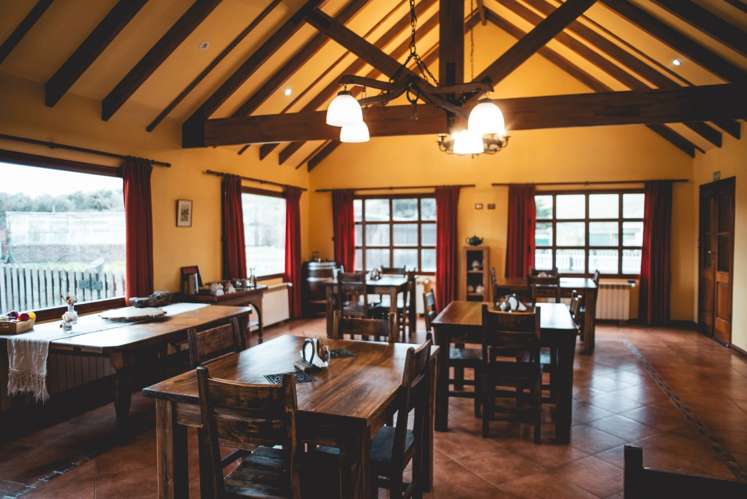 Sala de comedor con mesas de madera y sillas, ventanas grandes con cortinas rojas, paredes amarillas y techo con vigas de madera y lámparas colgantes.