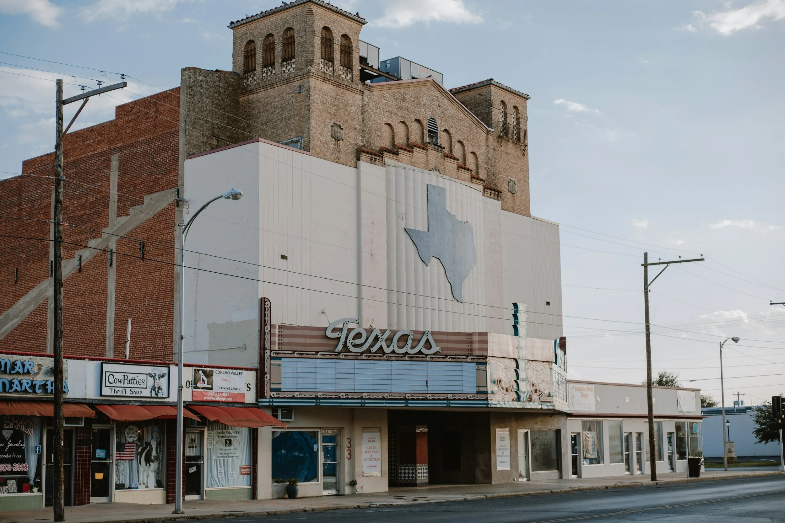 Street view of buildings with a large Texas sign and storefronts, including a thrift shop and other small businesses, under a cloudy sky.