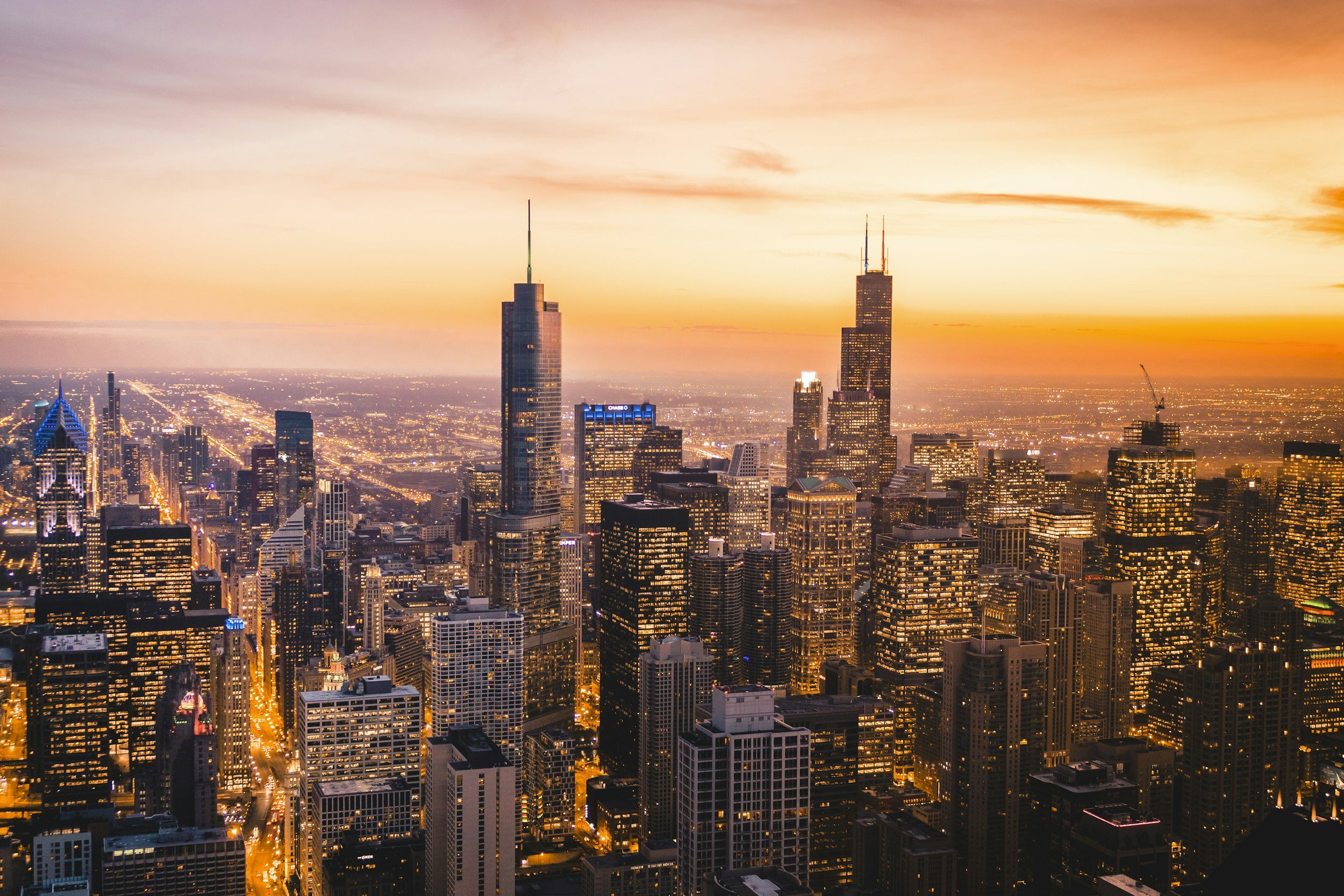 Aerial view of the Chicago skyline at sunset, featuring tall skyscrapers with lights beginning to turn on, and a colorful sky with orange and pink hues.