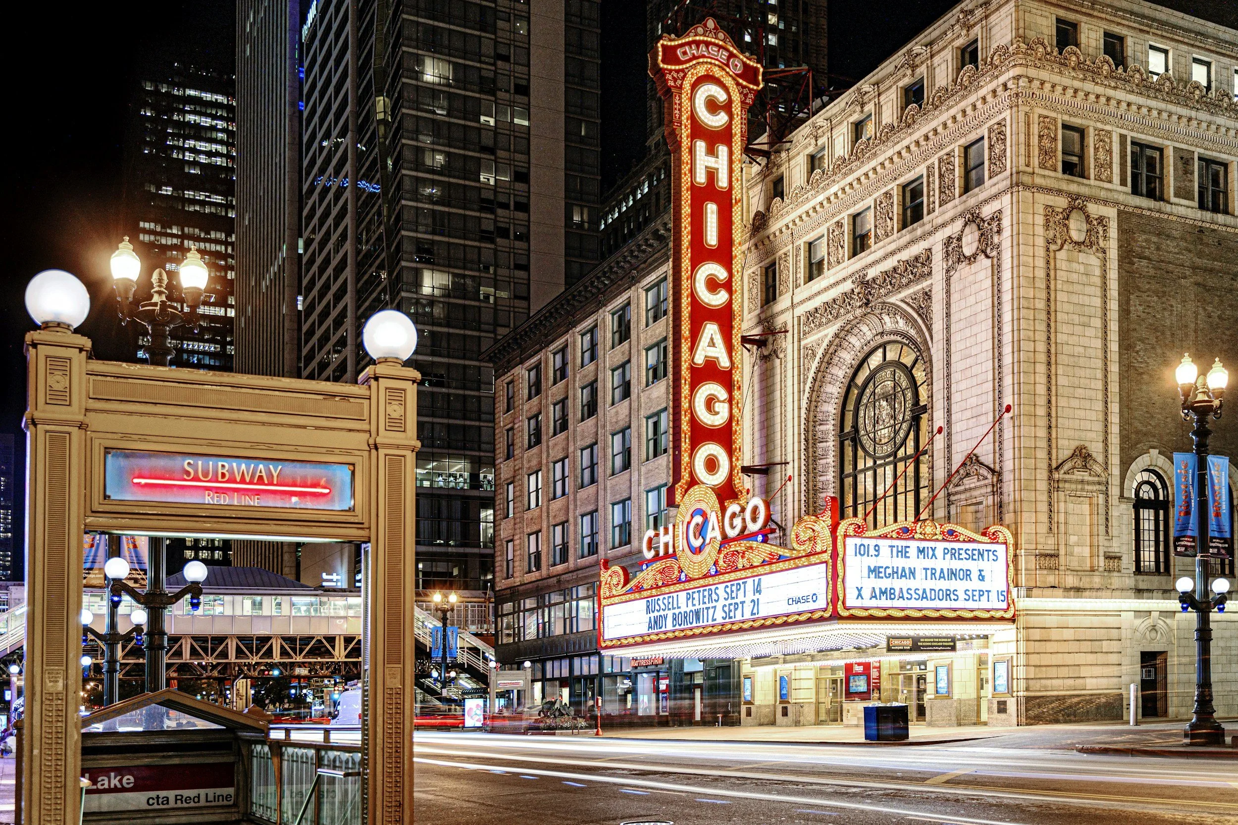 Nighttime scene of Chicago with neon theater signs, including a vertical 'ChicagO' marquee, a digital display with event listings, and streetlights illuminating the area.