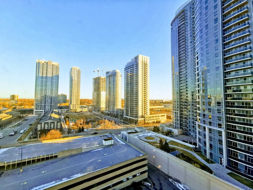 A cityscape with tall modern glass apartment buildings and office towers under a clear blue sky, with some trees and parking structures in the foreground.