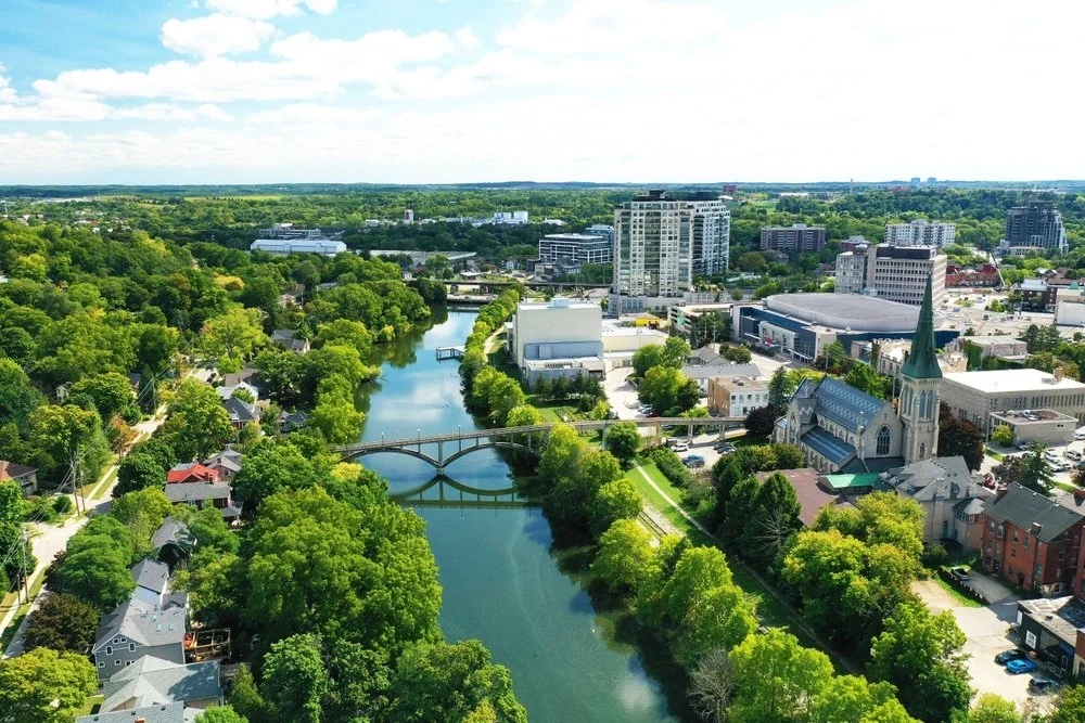 Aerial view of a city with a river running through it, surrounded by green trees and buildings, including a historic church with a tall steeple.